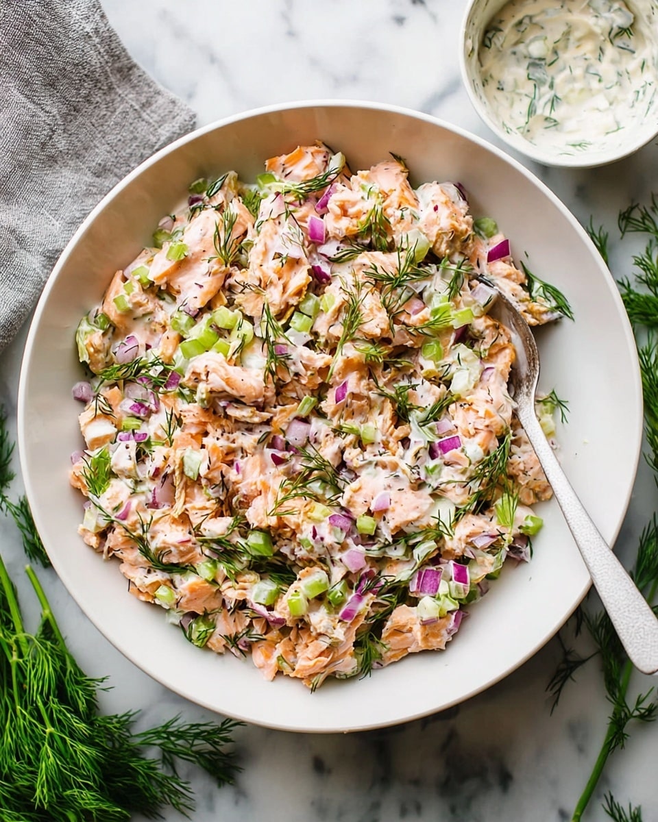 The image shows a white bowl filled with a mixed dish containing chunks of light orange fish pieces, small diced green celery, finely chopped red onion, and green herbs evenly spread throughout a creamy white sauce. Below the bowl, there is a white round plate with a thin brown rim, holding three open lettuce cups. Each cup is made from fresh green leaf lettuce and filled with the same fish and vegetable mix, showing all the textured colors in contrast with the bright green leaves. A silver spoon rests partly in the bowl on the right. The background is a white marbled surface. Photo taken with an iphone --ar 4:5 --v 7