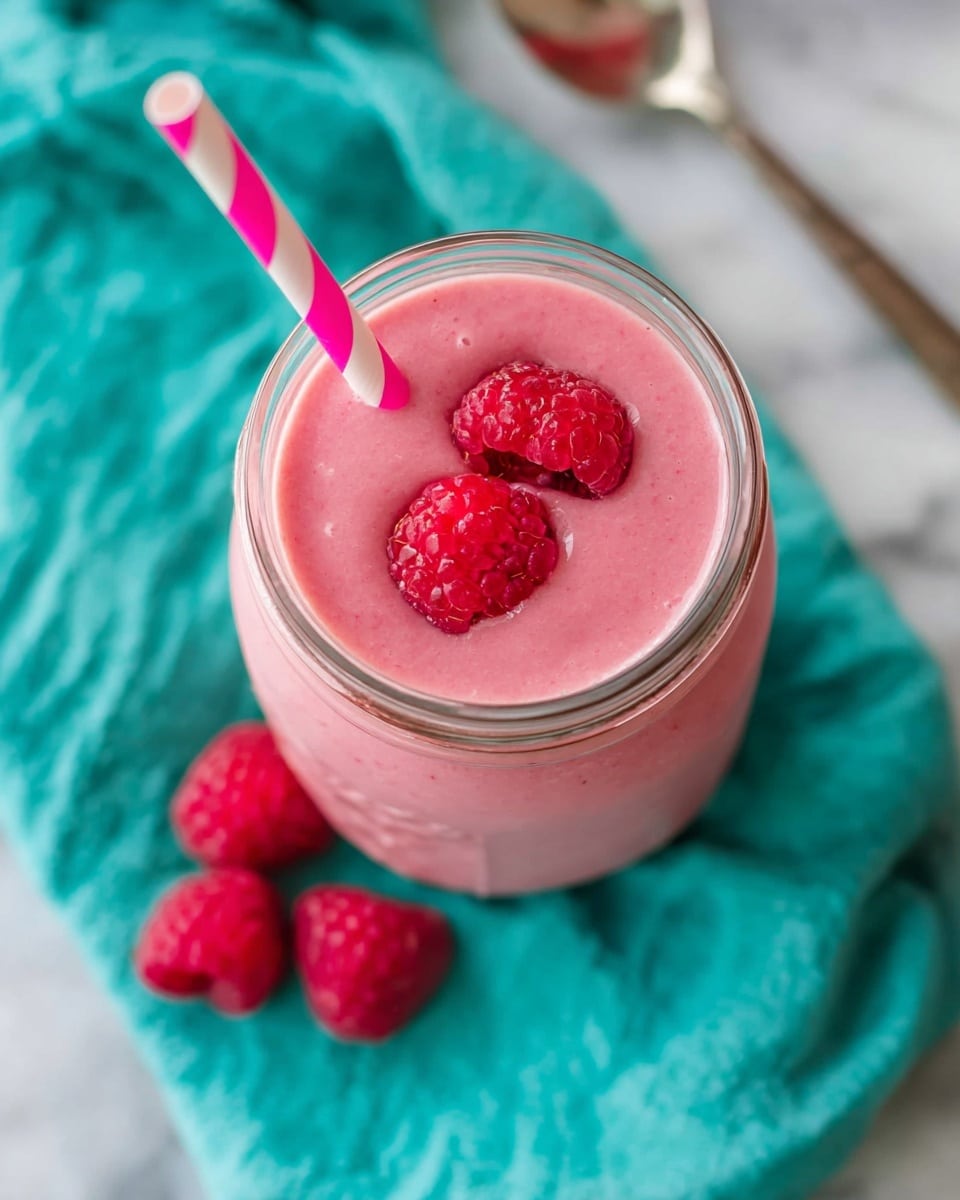 A tall clear glass jar filled with a pink raspberry smoothie showing a smooth, creamy texture. Two fresh red raspberries sit on top, slightly sinking into the smoothie. A pink and white striped straw is placed upright in the jar. The jar rests on a blue patterned cloth with some fresh raspberries scattered around and a knife with peanut butter on its blade next to the jar. The background is a white marbled surface. Photo taken with an iphone --ar 4:5 --v 7