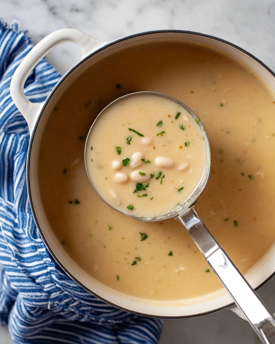 A close-up view of a white pot filled with thick, creamy beige soup that has small white beans visible throughout. The soup is lightly garnished with small, bright green chopped herbs scattered on top. A shiny silver ladle with a white handle is dipped into the soup, lifting a portion that shows the smooth texture of the soup with beans and herbs sitting on the surface. The pot is placed on a white marbled surface, and a blue and white striped cloth is partially visible beside the pot. Photo taken with an iphone --ar 4:5 --v 7