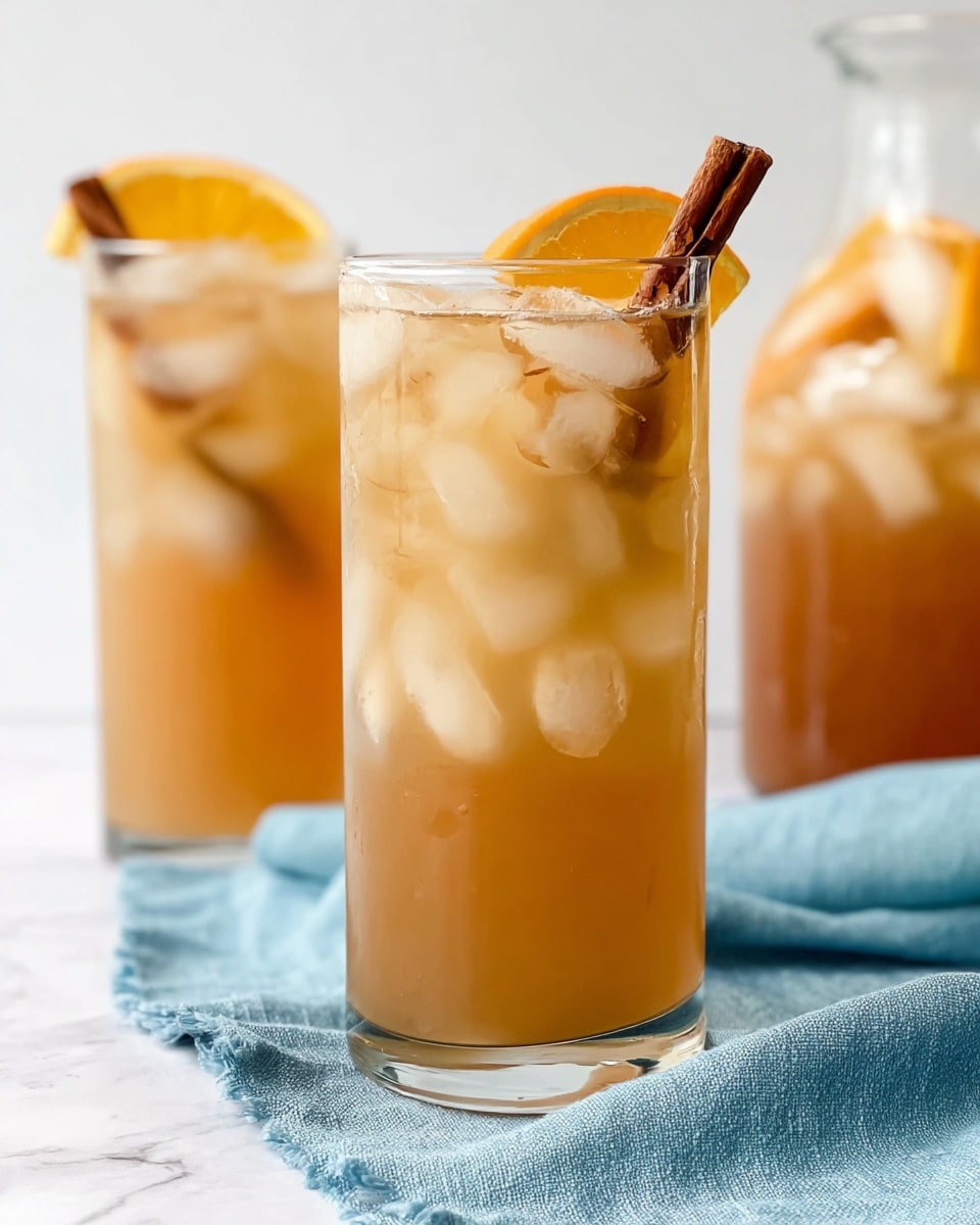 A clear glass pitcher filled with a light brown drink is shown; inside the pitcher are layers of floating fruit slices including a round orange slice near the top, a pale yellow apple slice above it, and a cinnamon stick standing upright among the liquid. The pitcher has a curved handle on the right and a spout with a small notch on the left. Around the base of the pitcher, there are whole apples and oranges, with one apple cut in half showing its white interior and seeds. The pitcher and fruits rest on a white marbled surface with a soft blue cloth partially visible behind it. Photo taken with an iphone --ar 4:5 --v 7