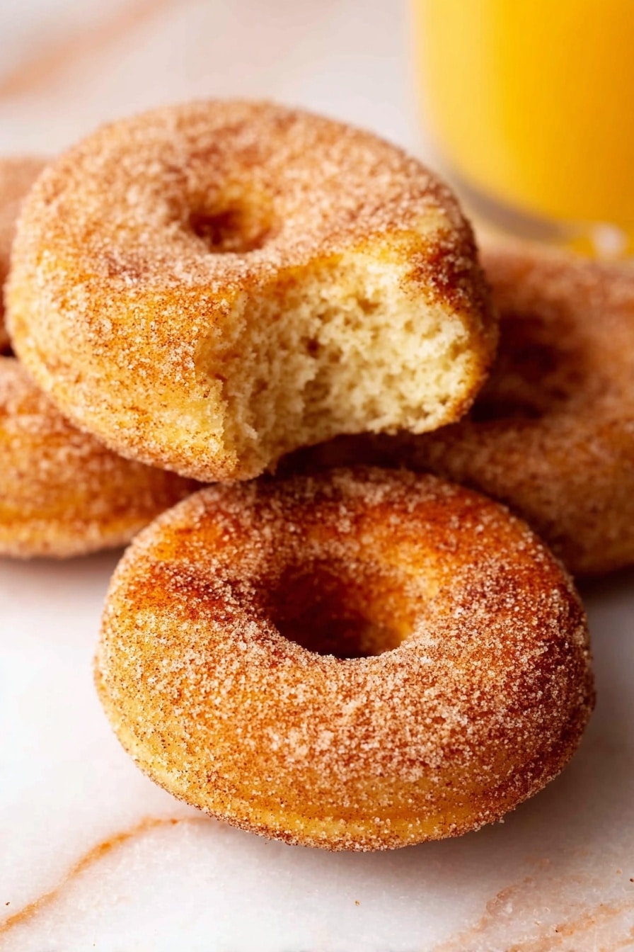 Vegan Apple Cider Donuts, fall vegan donut recipes, baked apple cider donuts, easy vegan fall treats, gluten-free vegan donuts - The image shows three cinnamon sugar donuts with a light golden brown color and a textured coating of sugar and cinnamon. One donut is in the front with a bite taken out, revealing a soft, fluffy inside with a slightly crumbly texture. The other two donuts are whole and placed closely behind it on a light-colored surface. The background has a white marbled texture, and there is a glass of orange juice visible on the right side of the image. photo taken with an iphone --ar 2:3 --v 7