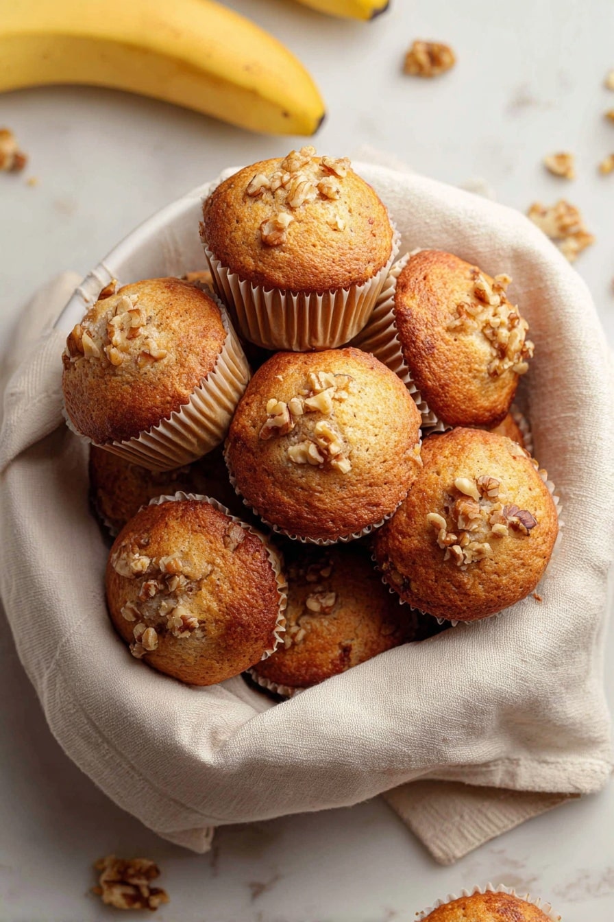 Quick & Easy Vegan Banana Muffins, vegan banana muffin recipe, healthy banana muffins, simple vegan muffin recipe, gluten-free banana muffins - A white bowl lined with a soft beige cloth holds a pile of golden brown muffins with slightly domed tops. Each muffin is in light beige paper cups and sprinkled with small pieces of walnuts on the top, giving a rough texture. Around the bowl on a white marbled surface, there are a few loose walnuts and bananas visible, adding natural color contrasts. The muffins appear moist with a lightly browned crust. photo taken with an iphone --ar 2:3 --v 7