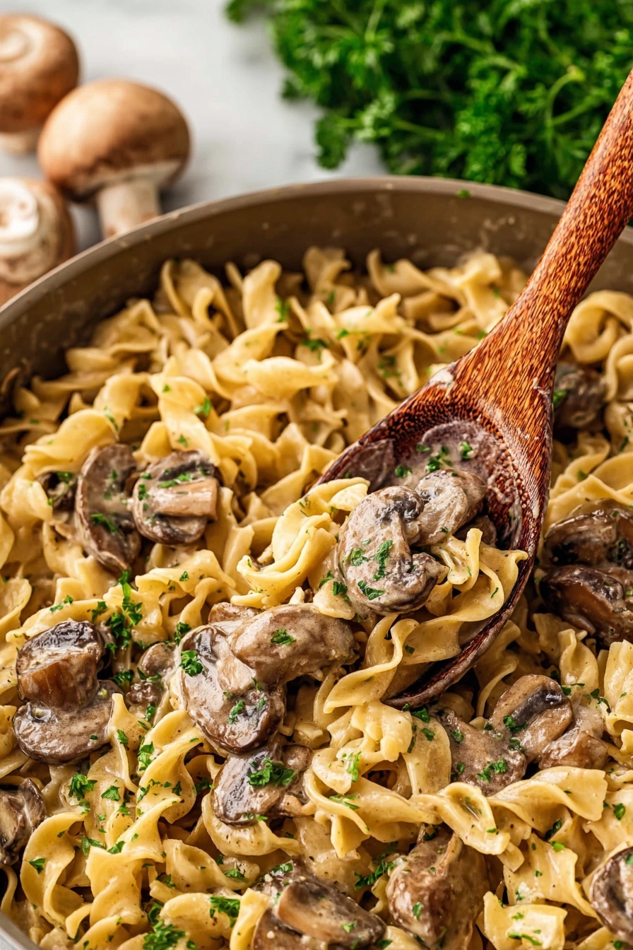 Old Fashioned Beef Stroganoff, beef stroganoff recipe, hearty beef stroganoff, classic beef stroganoff with mushrooms, creamy beef stroganoff - The image shows a close-up of a pan filled with a creamy noodle and mushroom dish. There are egg noodles that are light yellow, soft, and curly, spread all over the pan as the first layer. On top of the noodles, there are sliced mushrooms with a brown color and slightly shiny surface from the sauce. Mixed with the mushrooms and noodles, there are small bits of green parsley scattered for color. A wooden spoon is scooping some of the dish from the pan. The background has some blurred green parsley and whole mushrooms placed on a white marbled surface. photo taken with an iphone --ar 2:3 --v 7