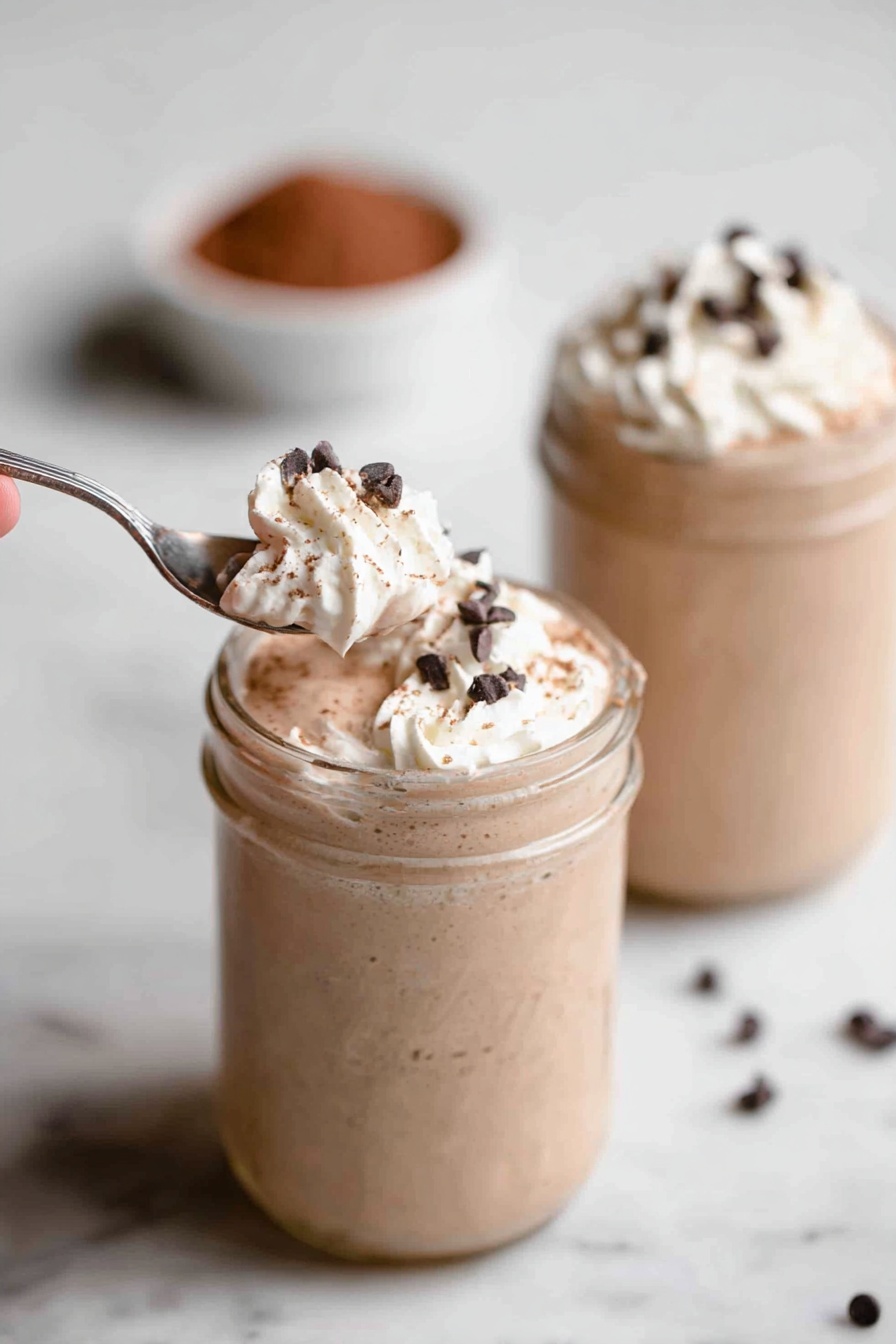 Vegan Chocolate Protein Shake, healthy vegan protein drinks, dairy-free chocolate shakes, plant-based protein smoothies, quick vegan breakfast recipes - Two glass jars sit on a white marbled surface, each filled with a creamy light brown milkshake. The milkshakes have a thick texture and are topped with a swirl of white whipped cream. Small, dark chocolate chips are sprinkled over the whipped cream. In the foreground, a silver spoon held by a woman's hand scoops a portion of whipped cream and chocolate chips from the first jar. The background jar is out of focus but shows the same whipped cream and chocolate chip topping. There is a small bowl filled with cocoa powder blurred in the background. Photo taken with an iphone --ar 2:3 --v 7