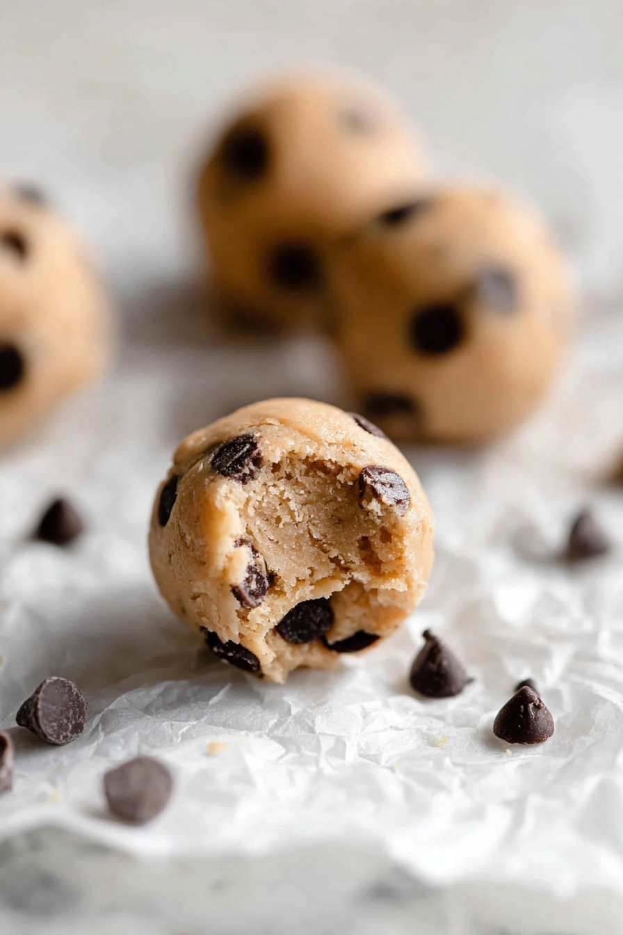 Protein Cookie Dough Bites, healthy no-bake snacks, high-protein dessert, easy protein bites, muscle-friendly treats - The image shows small round cookie dough balls with a light tan color, filled with many dark brown chocolate chips scattered inside and on the surface. The balls rest on a crumpled white paper on a white marbled background. One dough ball in the front has a small bite taken out of it, showing a soft, slightly crumbly texture inside, while other dough balls and some loose chocolate chips are blurred in the background. The lighting is soft and natural, highlighting the texture of the dough and chocolate chips, photo taken with an iphone --ar 2:3 --v 7