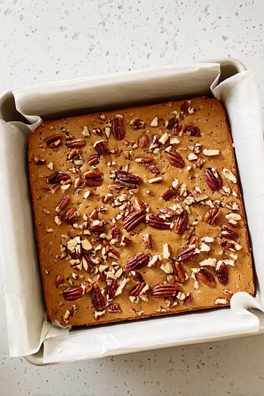 Maple Pecan Blondies, Maple Pecan Blondies Recipe, Easy Blondies with Maple and Pecans, Best Maple Pecan Blondies, Nutty Maple Blondies - A square baked dessert with a light brown top covered in scattered pieces of pecans. It is in a white baking pan lined with parchment paper that folds over the edges. The baked item looks soft and slightly puffy, with a smooth but slightly textured surface from the nuts. The pan sits on a white marbled surface. photo taken with an iphone --ar 2:3 --v 7