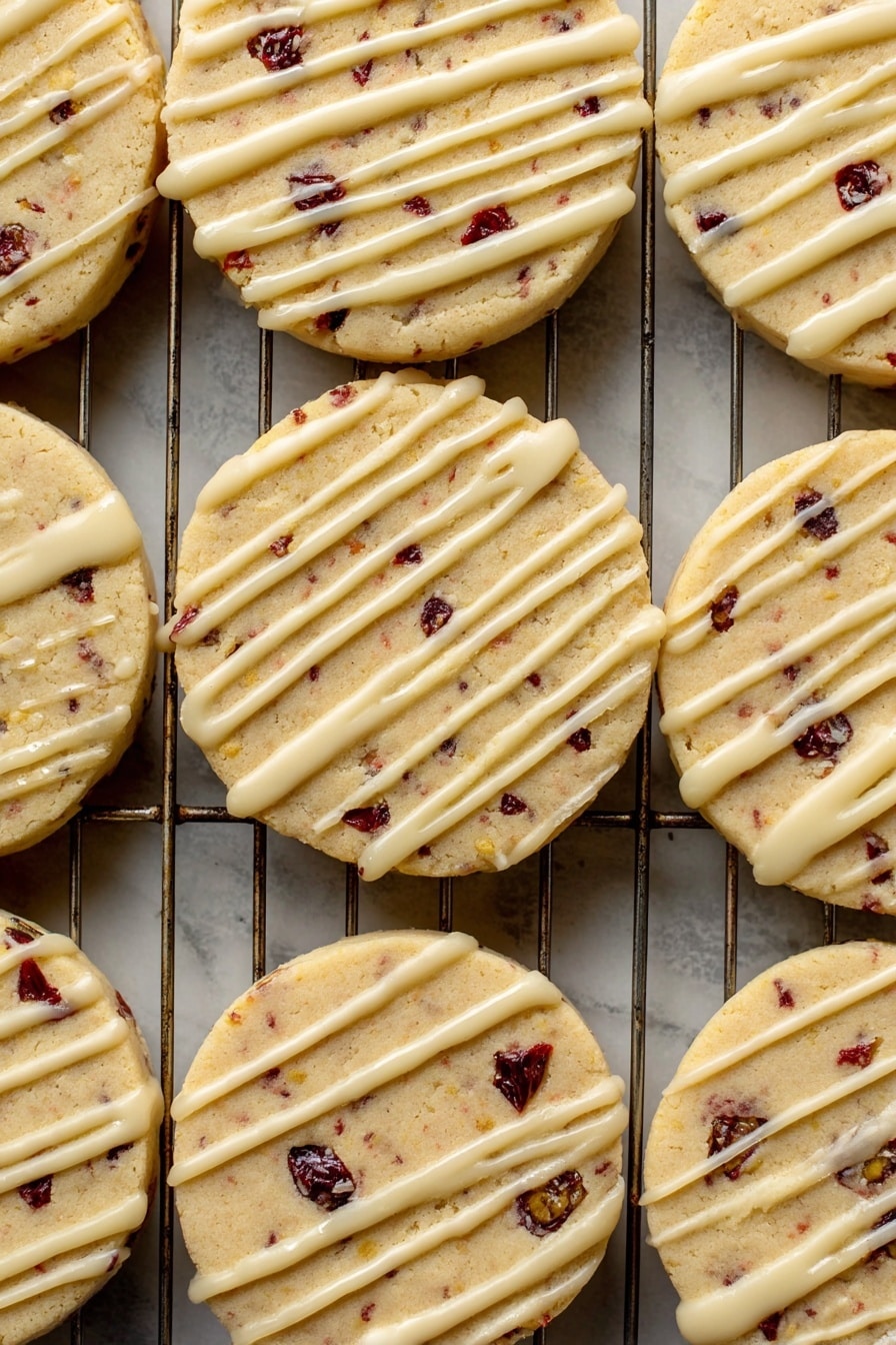 Orange Cranberry Shortbread Cookies, citrus cranberry cookie recipe, easy holiday shortbread, buttery cranberry cookies, festive citrus dessert - A close-up view of a batch of round cookies laid out on a metal cooling rack over a white marbled surface. Each cookie has one visible layer, light golden in color with small dark red and black fruit bits inside, giving a spotted look. On top of each cookie is a thin, even drizzle of pale cream-colored icing, applied unevenly in long lines across the surface. The texture of the cookies looks soft but firm, with some slight cracks near the edges. The metal wires of the rack create a grid pattern underneath the cookies. Photo taken with an iphone --ar 2:3 --v 7