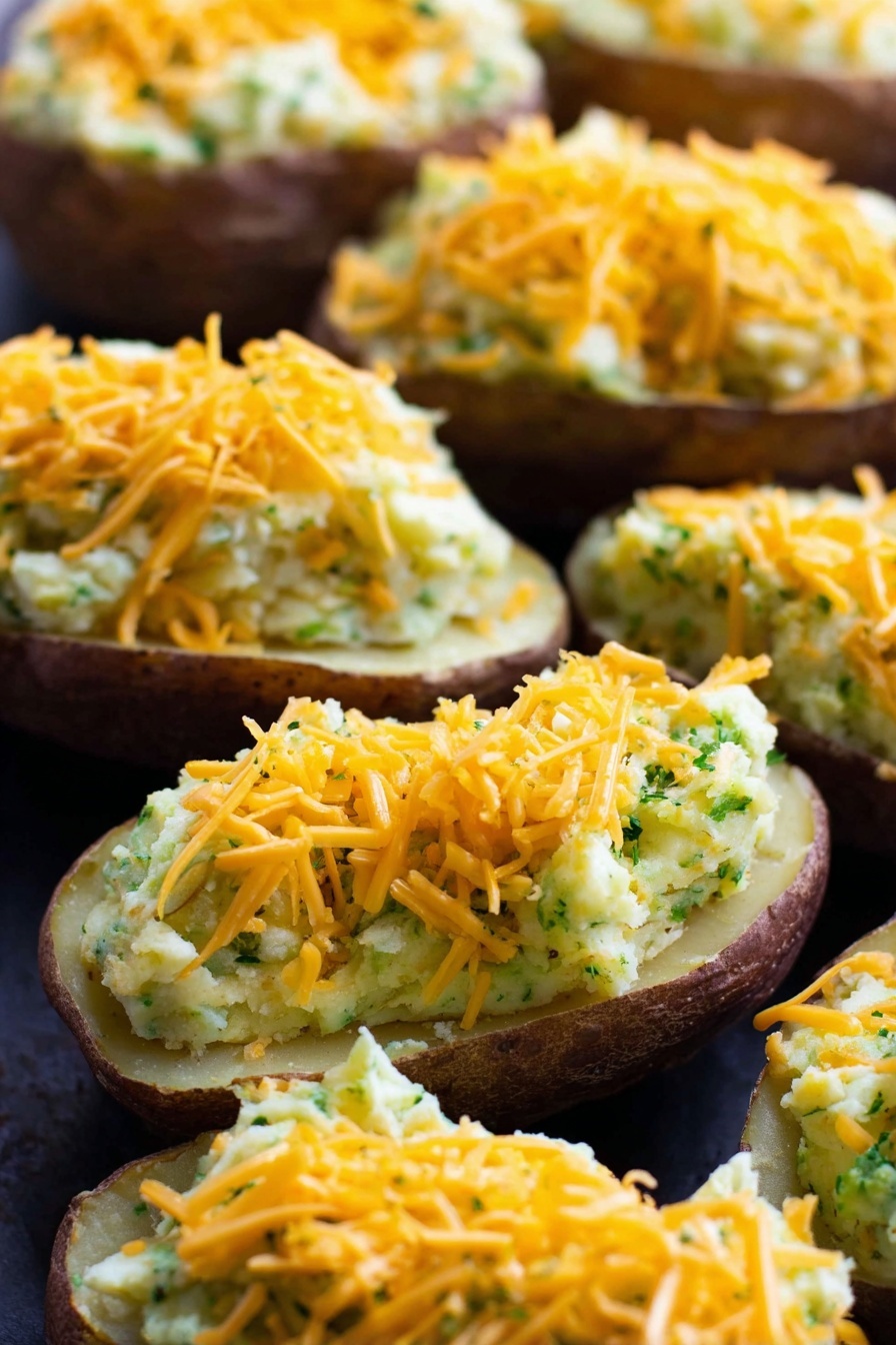 Vegan Cheesy Broccoli Twice Baked Potatoes, vegan loaded baked potatoes, dairy-free cheesy potatoes, healthy vegan stuffed potatoes, vegan comfort foods - The image shows several potato halves with the inside scooped out and filled with a creamy mixture that looks light yellow with green bits throughout, suggesting herbs or vegetables mixed in. Each stuffed potato is topped with a generous layer of shredded orange cheddar cheese. The potatoes are arranged closely together on a dark surface which contrasts with the light filling and bright cheese. The texture of the filling appears smooth but slightly chunky, and the potato skins are a dark brown color. The close-up shot highlights the layers clearly with the cheese on top, the creamy filling in the middle, and the potato skin at the bottom. Photo taken with an iphone --ar 2:3 --v 7