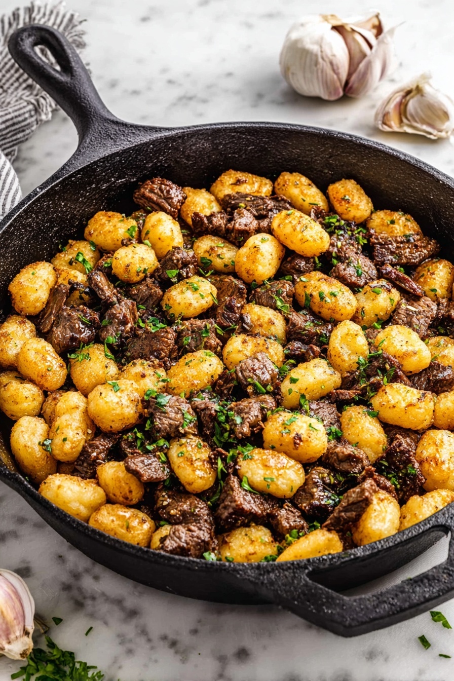 Steak Bites with Gnocchi in Garlic Butter, steak bites recipe, garlic butter gnocchi, easy steak and gnocchi dish, savory beef gnocchi - A cast iron pan is filled with two main layers: the first layer is browned small gnocchi pieces that are golden with a slightly crisp texture, and the second layer is tender, dark brown bite-sized beef chunks mixed evenly with the gnocchi. Small green parsley pieces are scattered on top, adding a touch of freshness and color contrast. The pan is sitting on a white marbled surface, with a few garlic bulbs placed nearby. The image is bright and clear showing the food textures well. photo taken with an iphone --ar 2:3 --v 7