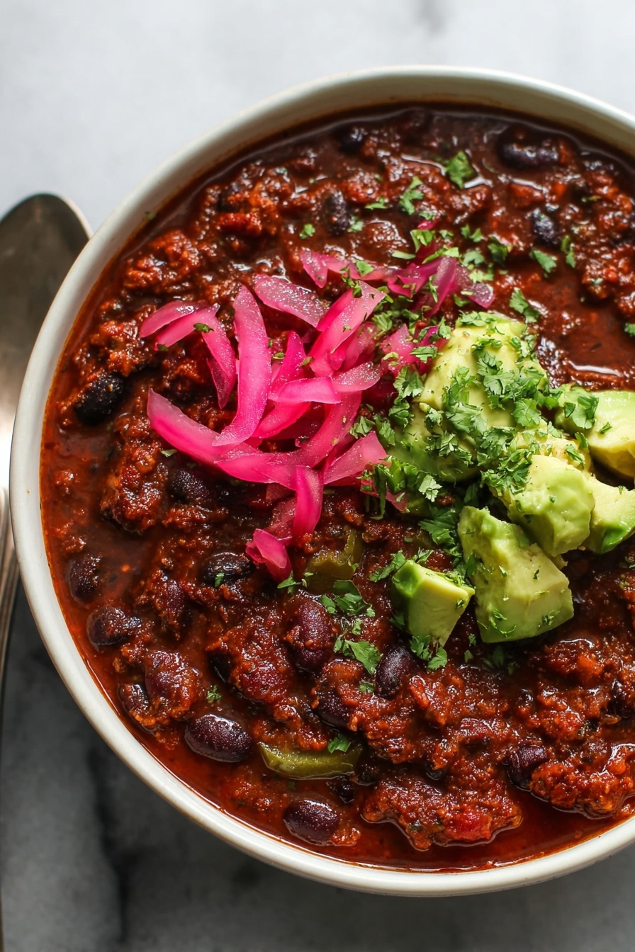 Lentil Chili, hearty vegan chili, healthy lentil recipes, vegetarian chili ideas, comforting chili with lentils - A close-up view of a bowl of thick chili, showing three main layers: a base layer of rich, dark red-brown chili with black and red beans and small pieces of green bell pepper spread throughout, topped on one side with bright pink pickled onion slices, and on the same side, fresh green avocado chunks sprinkled with finely chopped cilantro. The bowl is white and a spoon is partially visible on the left side, all placed on a white marbled surface. Photo taken with an iphone --ar 2:3 --v 7