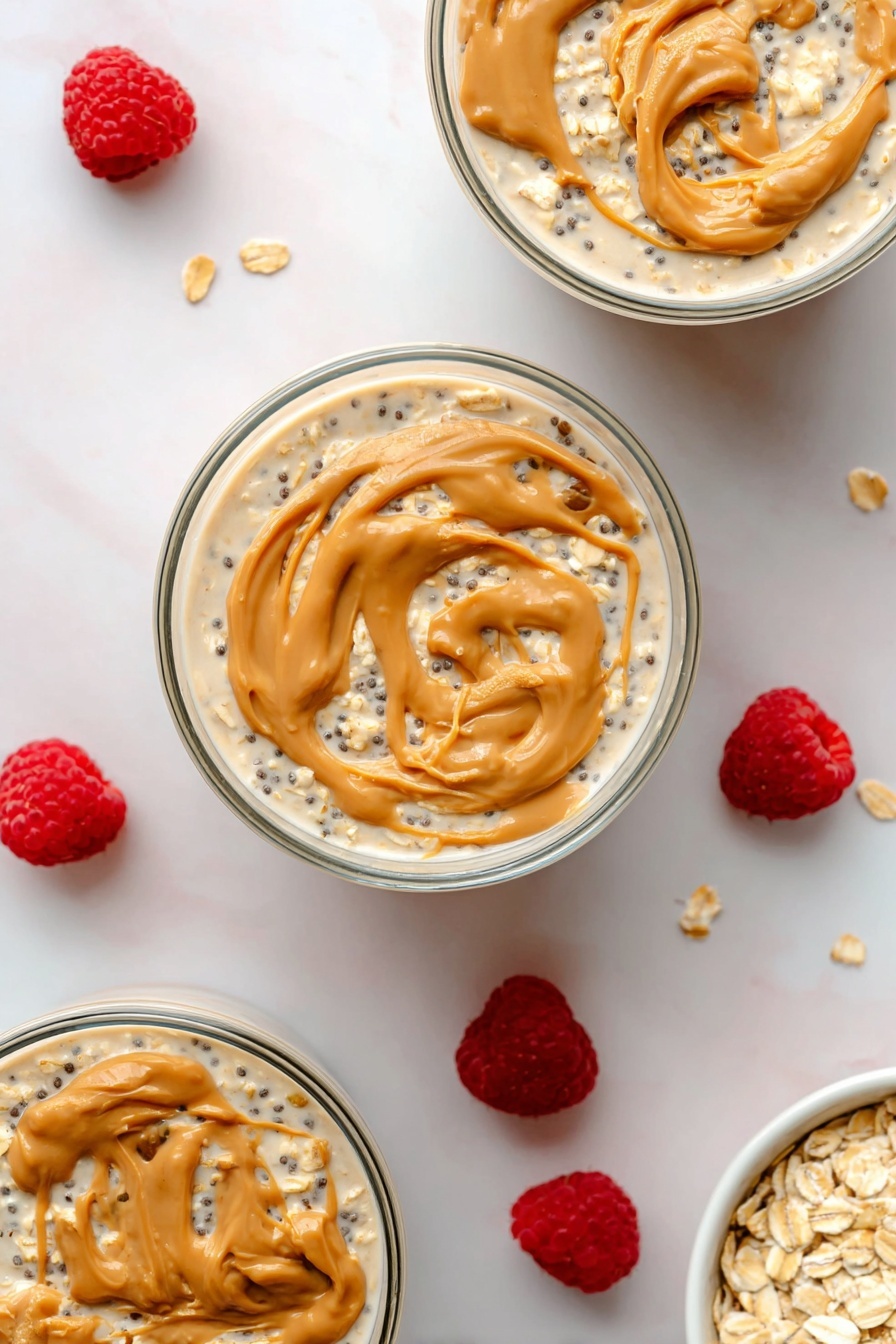 Peanut Butter Overnight Oats, healthy breakfast ideas, quick overnight oats, easy make-ahead breakfast, nutritious oatmeal recipes - The image shows three round clear glass bowls filled with layered overnight oats on a white marbled surface. Each bowl has a bottom layer of creamy light beige oats mixed with small black chia seeds, covered by a thin layer of milky liquid. The top layer is swirled with dollops of light brown peanut butter, creating smooth, curved textures on the surface. Around the bowls, scattered fresh bright red raspberries add a pop of color, along with a small white bowl filled with rolled oats placed in the bottom right corner. The photo taken with an iphone --ar 2:3 --v 7