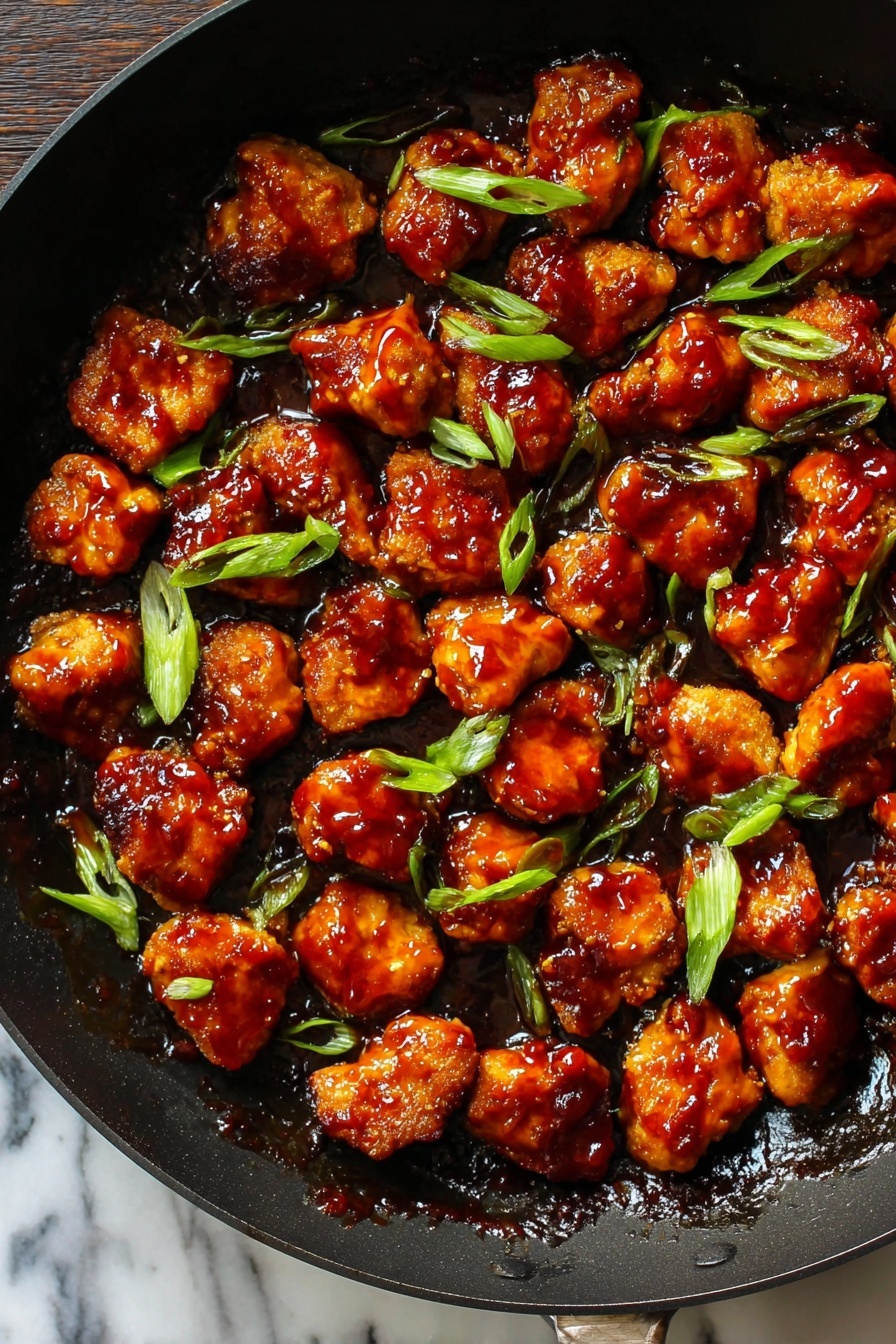 Honey Ginger Seitan with Crispy Pan-Fried Glaze, plant-based honey ginger seitan, vegan crispy stir-fry, easy seitan recipes, flavorful plant-based dishes - This image shows a black pan filled with many small pieces of golden-brown chicken covered in thick, shiny reddish-brown sauce. The pieces are spread out evenly across the pan, with some bright green slices of scallion sprinkled on top. The sauce looks sticky and glossy, covering all the chicken pieces in a rich layer. The pan's dark color contrasts with the vibrant colors of the chicken and scallions. The background surface is a white marbled texture. photo taken with an iphone --ar 2:3 --v 7