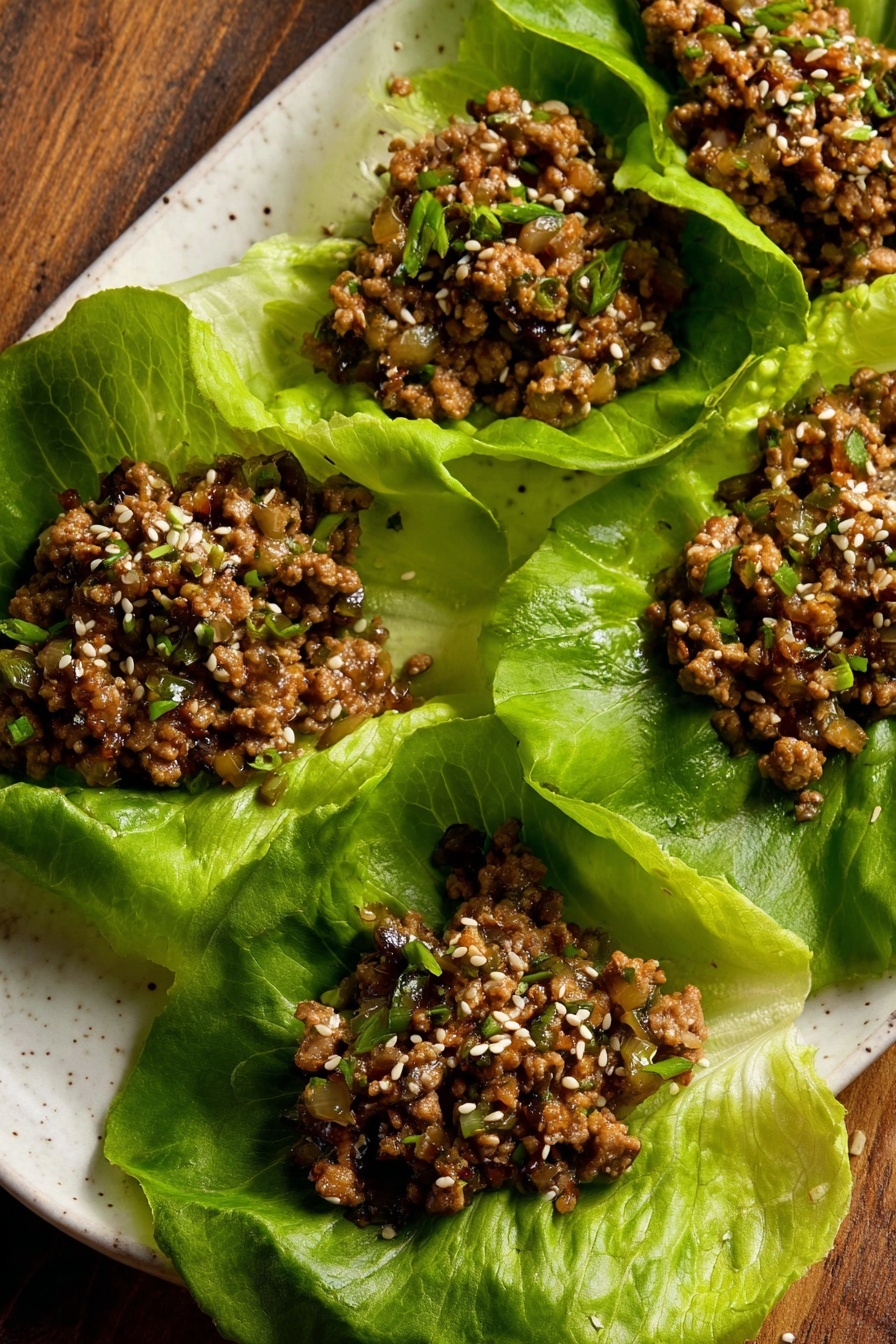 Tofu Lettuce Wraps, vegetarian lettuce wraps, healthy tofu stir-fry, vegan lettuce wraps, easy tofu lettuce wraps - The image shows five large green lettuce leaves arranged on a white plate with a wood surface in the background. Each leaf holds a serving of cooked ground meat mixed with small diced vegetables, with a brown color and a slightly moist texture. The meat mixture is sprinkled with white sesame seeds and small bits of green herbs. The lettuce leaves are fresh with visible veins and crinkled edges, creating a bright green contrast to the darker meat filling. Photo taken with an iphone --ar 2:3 --v 7