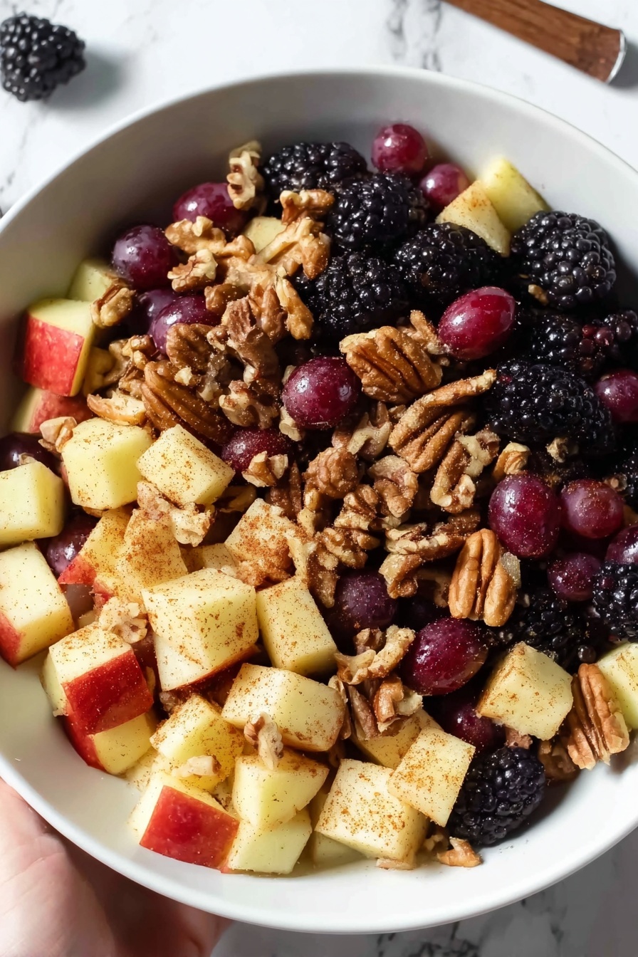 Fall Fruit Salad with Orange Maple Cinnamon Dressing, fall fruit salad, orange maple cinnamon dressing, seasonal fruit salad, easy fall fruit salad - A white bowl is filled with a fruit salad that has three main layers: the bottom layer is made of blackberries, which are bumpy and dark purple-black; the middle layer has round red grapes with smooth, shiny skin; and the top layer contains apple chunks that are pale yellow with a dusting of brown cinnamon powder. Scattered throughout the salad are light brown pecans adding a rough texture. The bowl sits on a white marbled surface, and two cinnamon sticks lie nearby. photo taken with an iphone --ar 2:3 --v 7