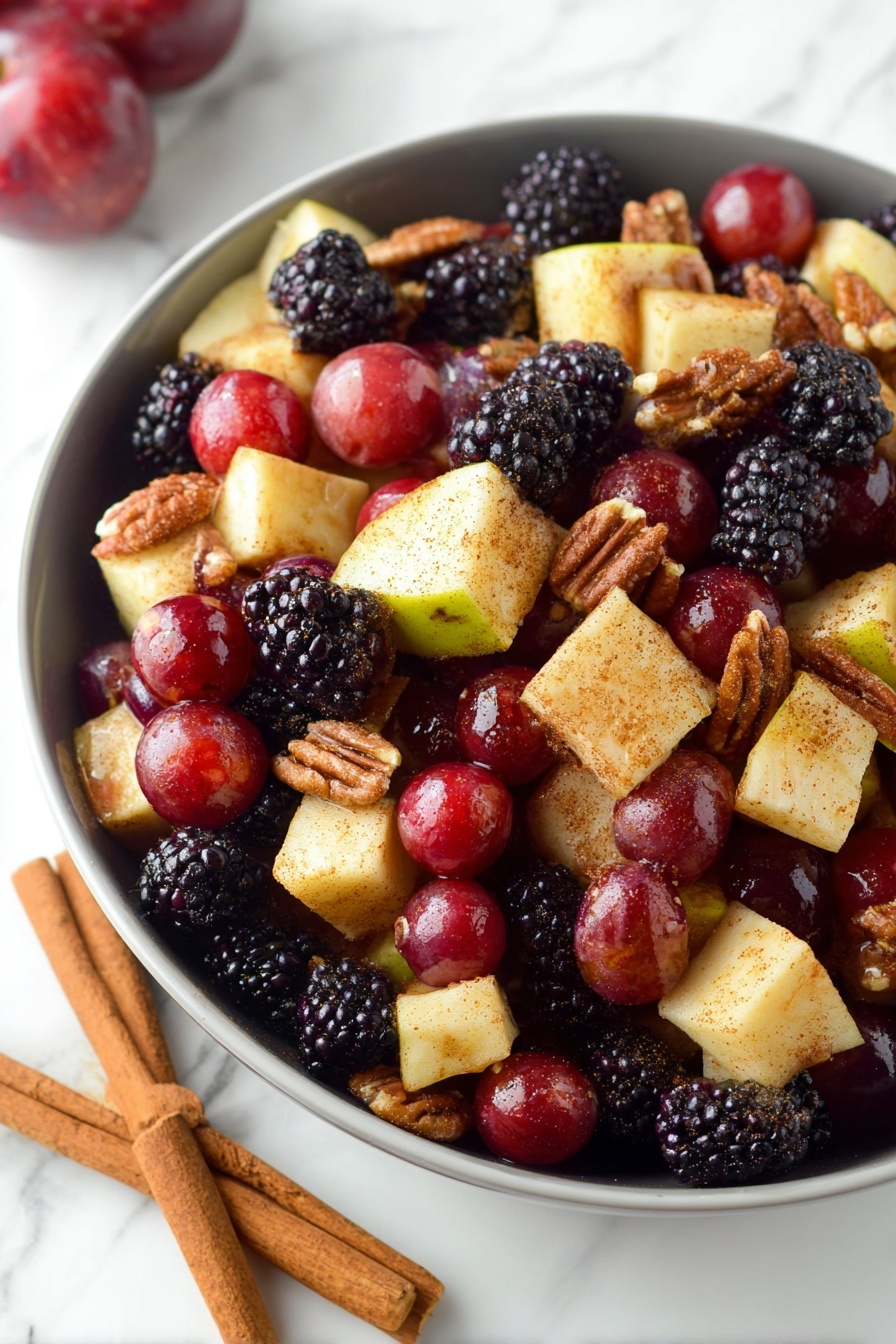 Fall Fruit Salad with Orange Maple Cinnamon Dressing, fall fruit salad, orange maple cinnamon dressing, seasonal fruit salad, easy fall fruit salad - A white bowl filled with a colorful fruit and nut salad, showing three layers of ingredients. The bottom layer is pieces of pale yellow apple with red skin, cut into small cubes. The middle layer features deep red grapes and dark purple-black blackberries scattered evenly across. The top layer includes crunchy brown walnuts and pecans mixed throughout. The fruit pieces have a light dusting of cinnamon or spice, giving a slight matte texture. The bowl is placed on a white marbled surface, and a woman's hand is visible holding the bowl's edge. photo taken with an iphone --ar 2:3 --v 7
