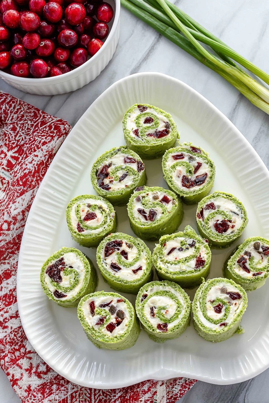 Cranberry Feta Pinwheels, Cranberry Feta Wraps, Easy Holiday Appetizer, No-Bake Cranberry Pinwheels, Festive Cheese Appetizer - On a white oval plate with a ridged edge, there are fifteen green pinwheel rolls arranged in a triangular shape, with one at the top, three in the second row, five in the third row, and six in the bottom row. Each roll shows a swirl of bright green outer layer with a creamy white filling inside that contains small pieces of dark red and green ingredients mixed throughout. The plate is placed on a white marbled surface, and to the top left, a white bowl filled with red berries is partially visible, sitting on a red and white patterned cloth. A few green onion stems lie on the top right corner of the surface. Photo taken with an iphone --ar 2:3 --v 7