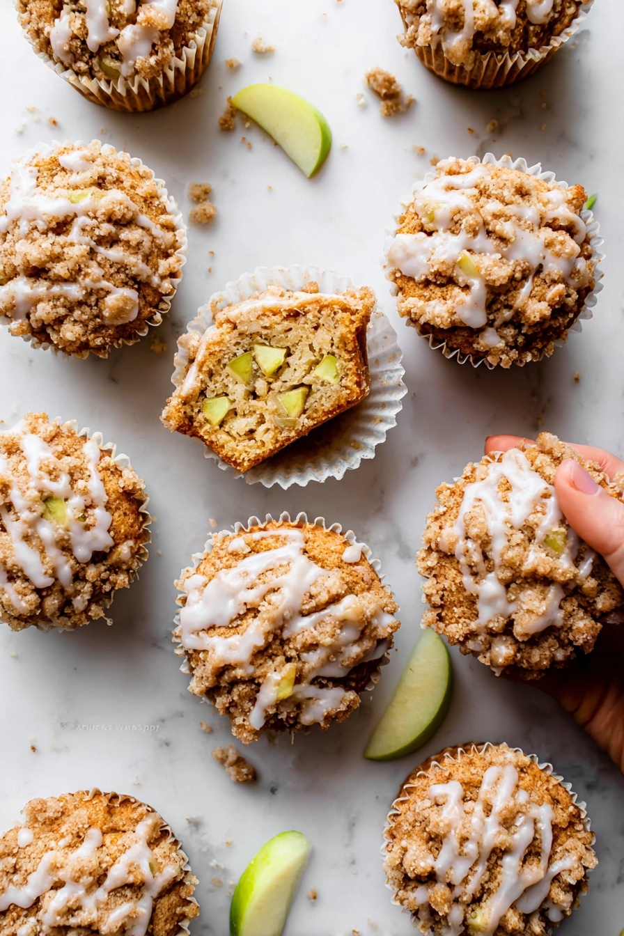 Apple Cinnamon Crumb Muffins, apple cinnamon muffins, cinnamon apple muffins, apple crumb muffins, fall breakfast muffins - The image shows nine apple crumb muffins arranged on a white marbled surface. Each muffin has three clear layers: a bottom baked golden-brown cake layer, a middle layer with visible light green apple chunks inside the muffin, and a top crumbly light brown streusel topping with a slight sprinkle of white glaze drizzled unevenly over it. One muffin is open, showing the inside texture and apple pieces, sitting in a white paper liner with a woman's hand about to pick it up. Small crumbs and a few green apple slices are scattered around, adding detail to the scene. Photo taken with an iphone --ar 2:3 --v 7