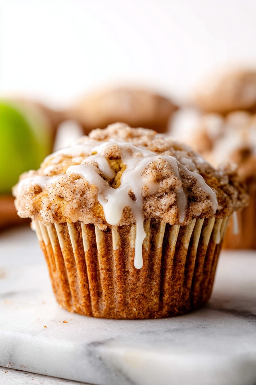 Apple Cinnamon Crumb Muffins, apple cinnamon muffins, cinnamon apple muffins, apple crumb muffins, fall breakfast muffins - A close-up of a single muffin on a white marbled surface, wrapped in a beige cupcake liner that is slightly opened, showing the muffin's ridged, golden-brown sides. The muffin has a crumbly top layer with a mix of light and darker brown textures and a smooth white glaze dripping down from the top, creating small streams over the crumb topping. In the background, there are blurred parts of other muffins and a hint of a green fruit, all against a bright white background. Photo taken with an iphone --ar 2:3 --v 7
