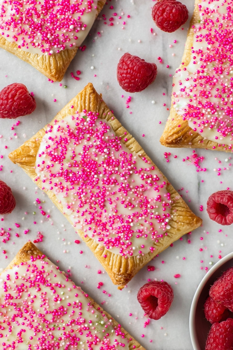 Homemade Raspberry Pop Tarts, raspberry pop tarts recipe, easy homemade pop tarts, flaky raspberry pastry, DIY raspberry breakfast treats - Several rectangular toasted pastries spread over a white marbled surface, each with a golden-brown crust stamped with fork marks along the edges; the top layer is coated in a slightly shiny white icing sprinkled generously with bright pink sugar crystals, creating a textured look. Scattered around the pastries are fresh, plump red raspberries adding a pop of natural color. In the bottom right corner, a small portion of a white bowl is visible, holding more raspberries. photo taken with an iphone --ar 2:3 --v 7