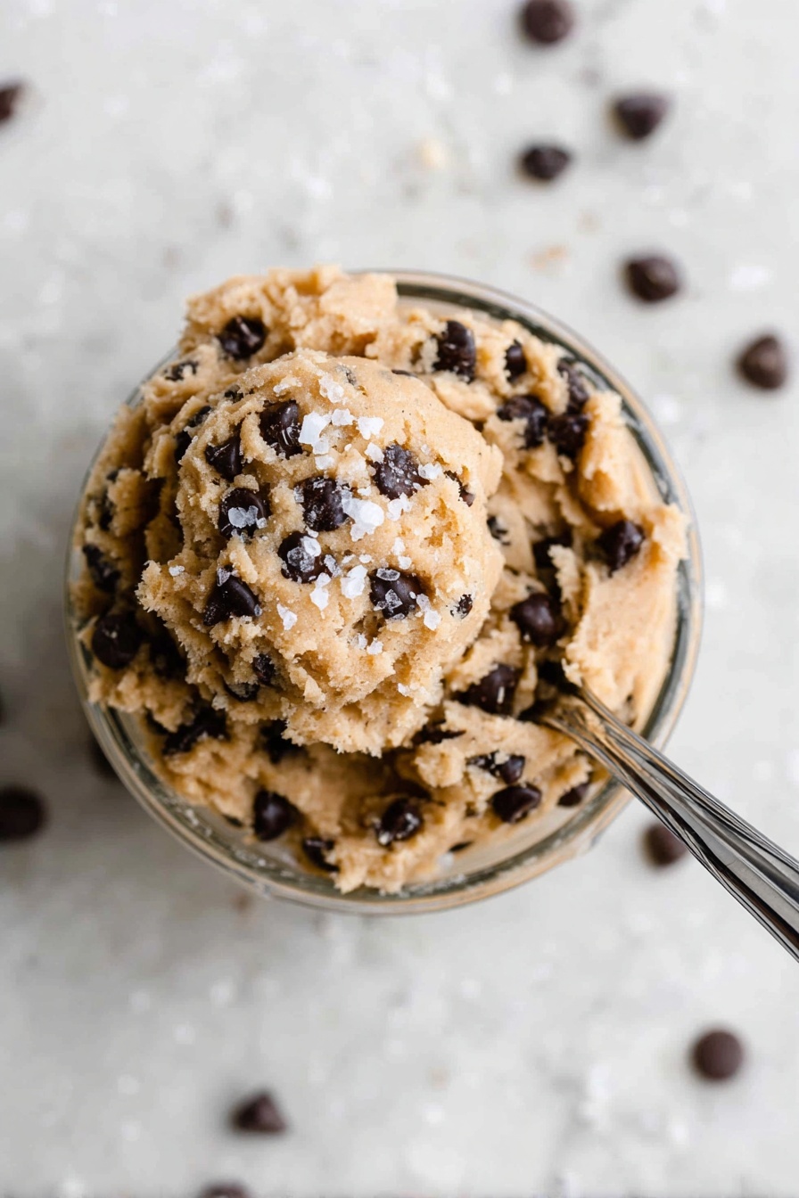 Protein Cookie Dough Bites, healthy no-bake snacks, high-protein dessert, easy protein bites, muscle-friendly treats - In a small clear glass bowl filled with one layer of light brown, soft cookie dough mixed with many small dark chocolate chips all over, the dough is slightly scooped at the top with a few coarse white salt flakes sprinkled on it. A shiny silver spoon is stuck into the dough from the right side, angled toward the top right corner, resting inside the bowl. The bowl sits on a surface with a white marbled texture, and some chocolate chips are scattered around it. The photo is taken with an iphone --ar 2:3 --v 7