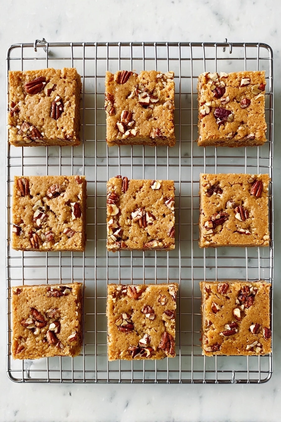 Maple Pecan Blondies, Maple Pecan Blondies Recipe, Easy Blondies with Maple and Pecans, Best Maple Pecan Blondies, Nutty Maple Blondies - Nine square pieces of light brown nut bars sit on a silver cooling rack. Each bar has small, uneven chunks of pecans scattered on top, creating a rough texture. The bars are evenly spaced in three rows and three columns, showing a soft, baked surface. The background is a white marbled texture. Photo taken with an iphone --ar 2:3 --v 7