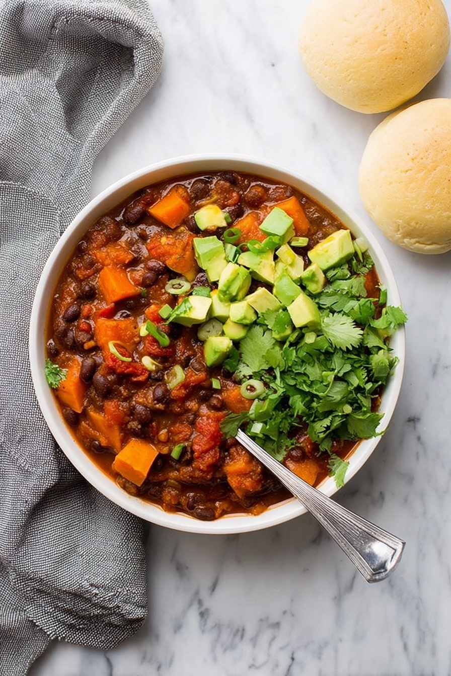 Sweet Potato Black Bean Chili, healthy vegetarian chili, hearty fall dinner, easy chili recipe, cozy plant-based meal - A white bowl filled with three main layers: the base is a thick stew of dark brown beans mixed with deep orange chunks of sweet potato and red tomato pieces in a reddish-brown sauce, topped on one side with bright green chopped avocado cubes and sprinkled with small rings of green onion, and on the other side with fresh green cilantro leaves. A silver spoon rests inside the bowl at the right side. The bowl sits on a white marbled surface next to two light beige round bread rolls and a loosely folded gray cloth. Photo taken with an iphone --ar 2:3 --v 7