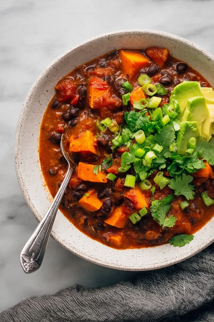Sweet Potato Black Bean Chili, healthy vegetarian chili, hearty fall dinner, easy chili recipe, cozy plant-based meal - The image shows a white speckled bowl filled with a thick stew made of dark black beans and bright orange chunks of sweet potato in a rich red tomato sauce. The stew is garnished with vibrant fresh green cilantro leaves and sliced green onions, while creamy green avocado pieces sit on one side near the bowl’s edge. A silver spoon is placed in the stew with its handle resting on the edge of the bowl. The bowl is set on a white marbled surface with a gray cloth draped nearby. photo taken with an iphone --ar 2:3 --v 7