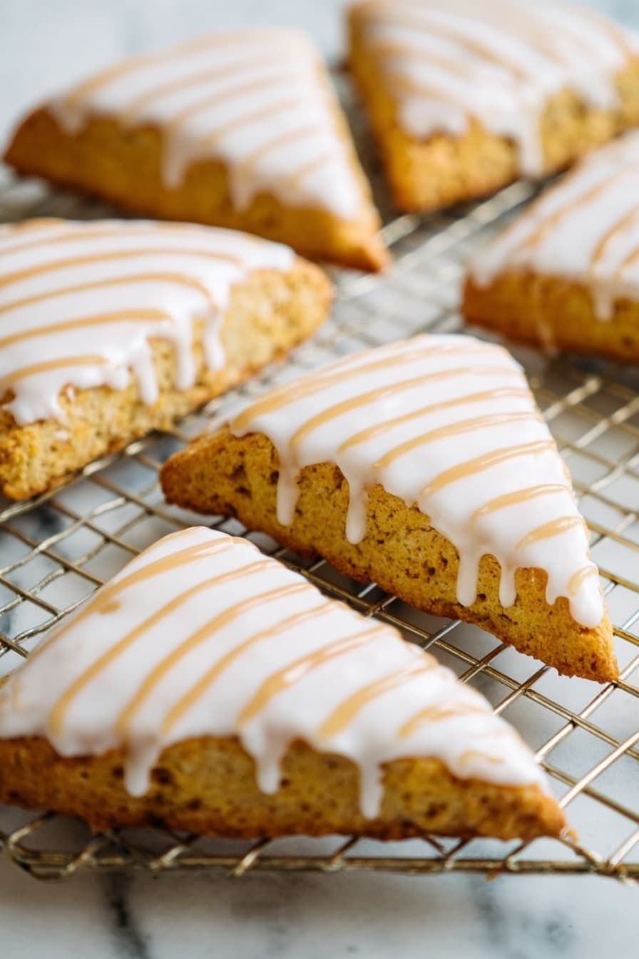Starbucks Copycat Vegan Pumpkin Scones, vegan pumpkin scones, plant-based pumpkin scones, gluten-free pumpkin scones, fall vegan baking ideas - The image shows a close-up of several triangular scones placed on a wire cooling rack over a white marbled surface. Each scone has a golden-brown, slightly rough texture base with a smooth, thick white icing layer covering the top. Over the white icing, there are thin, even lines of light brown drizzle arranged diagonally from one corner to the other. The scones are arranged in a casual pattern, filling the frame with their detailed textures and colorful toppings. photo taken with an iphone --ar 2:3 --v 7