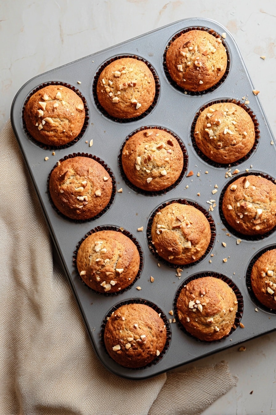Quick & Easy Vegan Banana Muffins, vegan banana muffin recipe, healthy banana muffins, simple vegan muffin recipe, gluten-free banana muffins - A grey muffin tray holds twelve freshly baked muffins, each with a golden-brown top that is slightly rounded and textured with small cracks. Light tan chopped nuts are scattered on the surface of each muffin, adding a crunchy detail. The muffins sit in dark brown paper liners that are barely visible around the edges. The tray rests on a white marbled texture surface with a soft beige cloth partially visible on the left side. The light gently highlights the warm tones and textures of the muffins, making them look soft and fresh. photo taken with an iphone --ar 2:3 --v 7