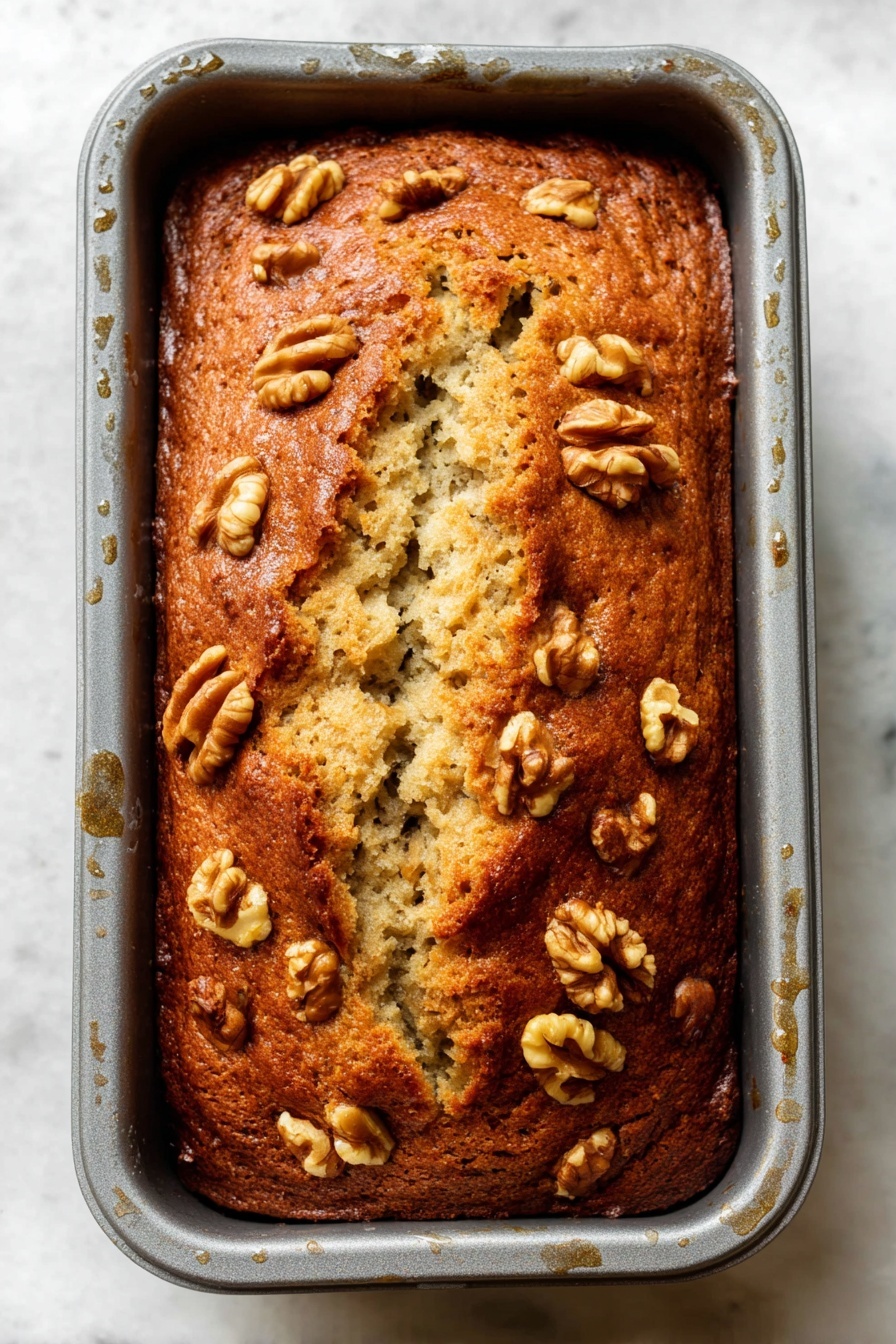 Best Vegan Banana Bread, vegan banana bread, egg-free banana bread, healthy banana bread, plant-based banana bread - A loaf of golden brown banana bread with a slightly cracked top sits inside a gray baking pan, showing the soft, light beige interior through the cracks. The surface is sprinkled with medium-sized walnut halves that are toasted to a light brown, scattered evenly on top and around the edges. The loaf has a dense, moist texture with visible bits of banana inside, and the baking pan has some visible moisture droplets on its sides. The background is a white marbled texture. Photo taken with an iphone --ar 2:3 --v 7