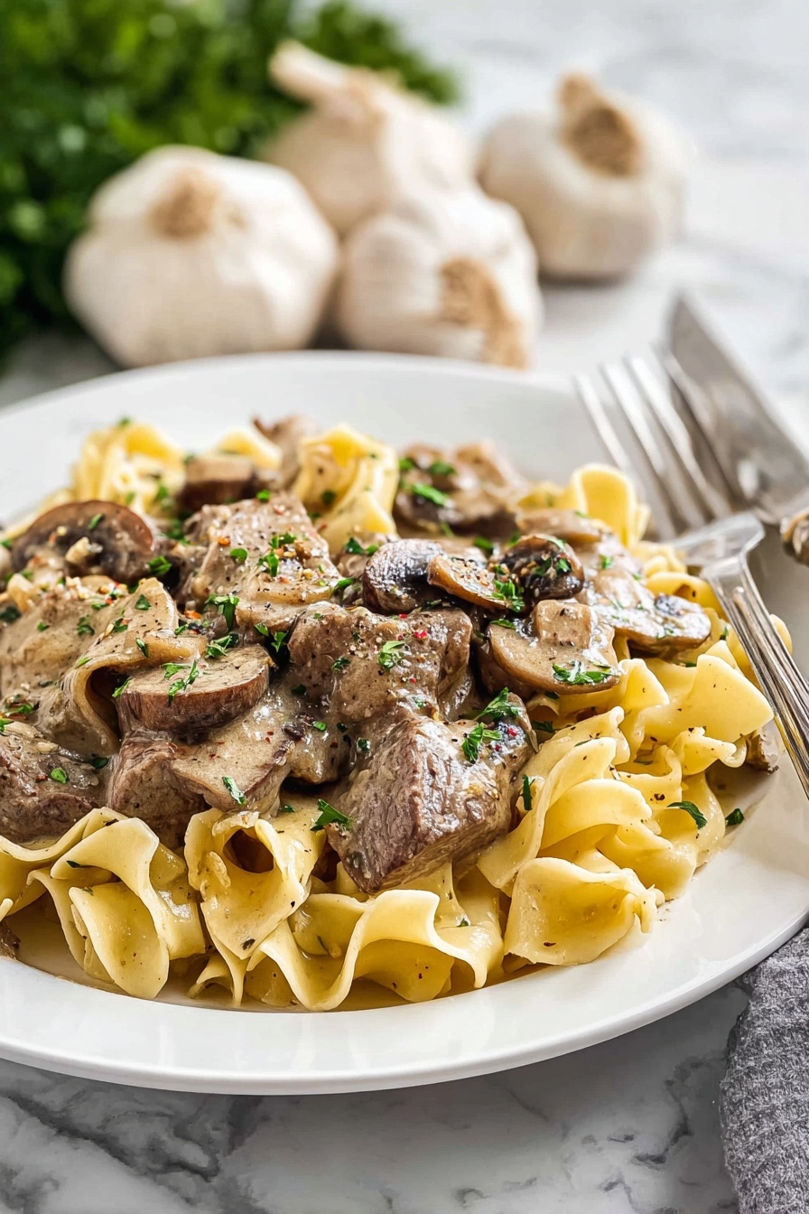 Old Fashioned Beef Stroganoff, beef stroganoff recipe, hearty beef stroganoff, classic beef stroganoff with mushrooms, creamy beef stroganoff - The image shows a close-up of a pan filled with creamy cooked egg noodles mixed with slices of brown mushrooms and small pieces of tender meat. The noodles are a light golden color with a soft texture, layered evenly throughout the pan. The meat and mushrooms are scattered on top and mixed in, adding shades of dark brown and gray. Bits of green herbs are sprinkled over the dish, adding some color contrast. A wooden spoon is resting in the pan, partially lifting a mix of noodles, mushrooms, and meat, suggesting that the dish is ready to be served. The background is a soft focus white marbled texture. Photo taken with an iphone --ar 2:3 --v 7