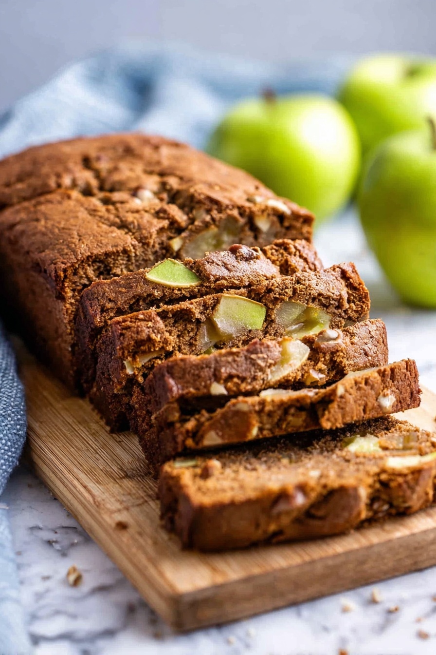 Moist Cinnamon Apple Bread, apple cinnamon loaf, fall breakfast bread, moist apple bread recipe, cinnamon apple cake - A loaf of brown apple bread with a soft, cracked crust sits on a wooden board placed on a white marbled surface. The bread is sliced into thick pieces, revealing a moist inside with chunks of green apple and nuts scattered throughout. The background has a few whole green apples and a light blue kitchen towel, slightly blurred to keep the focus on the bread. photo taken with an iphone --ar 2:3 --v 7