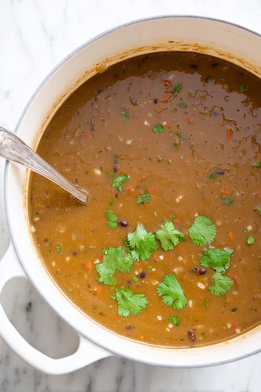 Vegan Black Bean Soup, vegan black bean soup recipe, healthy black bean soup, hearty vegan soup, easy vegan black bean dish - A large white pot filled with thick brown soup that has a slightly grainy texture, small visible pieces of vegetables, and beans mixed throughout. Several fresh green cilantro leaves float on top, adding a touch of bright green color. A silver spoon is partially dipped into the soup on the left side of the pot. The pot is placed on a white marbled surface. Photo taken with an iphone --ar 2:3 --v 7