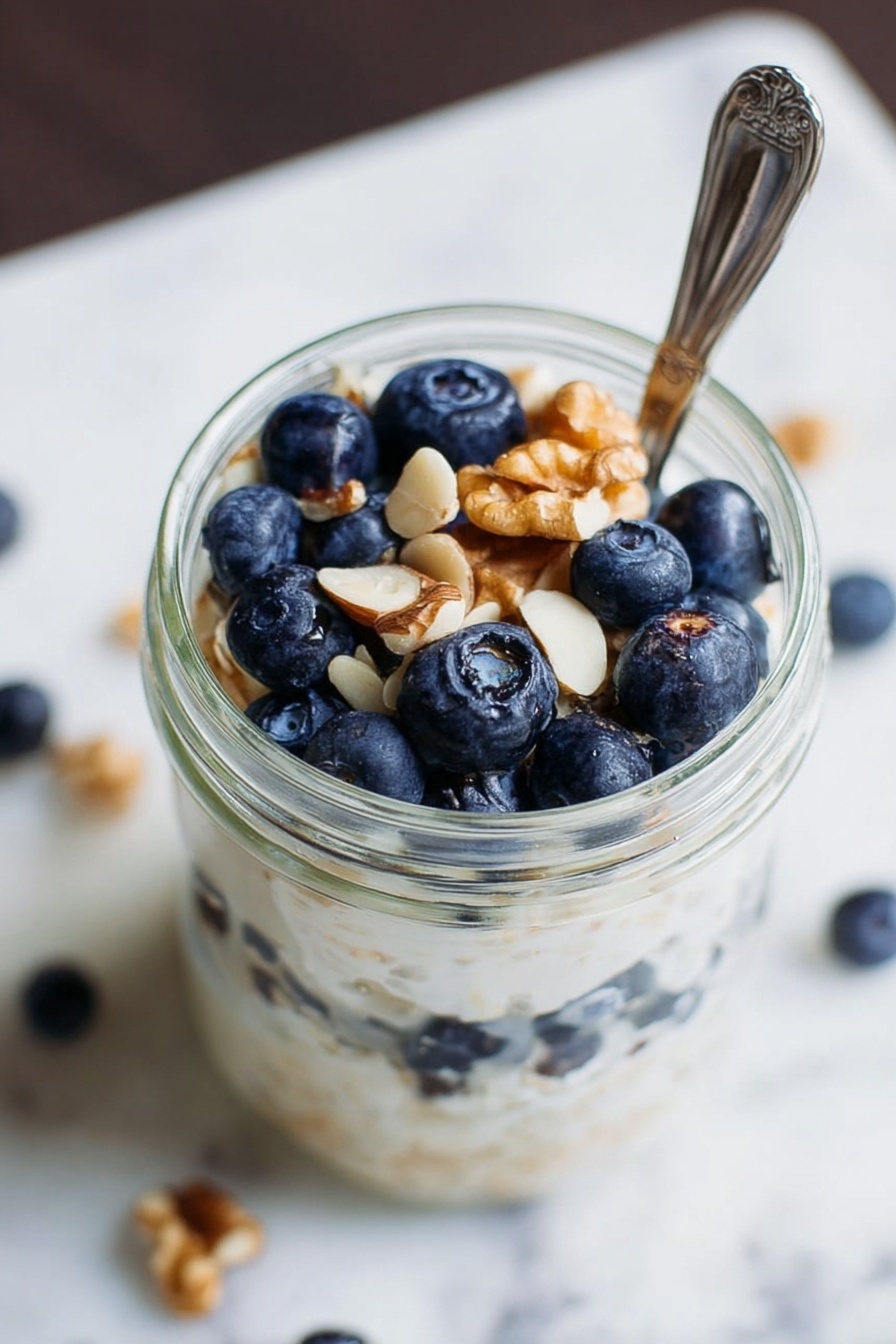 Simple Vegan Overnight Oats, vegan breakfast ideas, easy overnight oats, healthy vegan breakfast, quick vegan morning recipes - A clear glass jar filled with three layers: the bottom layer is fresh blueberries, the middle layer is creamy white porridge or yogurt with a smooth texture, and the top layer has more blueberries and a few light brown walnut pieces scattered on top. A silver spoon is placed in the jar, partly inside the food. The surface around the jar shows a white marbled texture with some scattered white nut pieces. Photo taken with an iphone --ar 2:3 --v 7