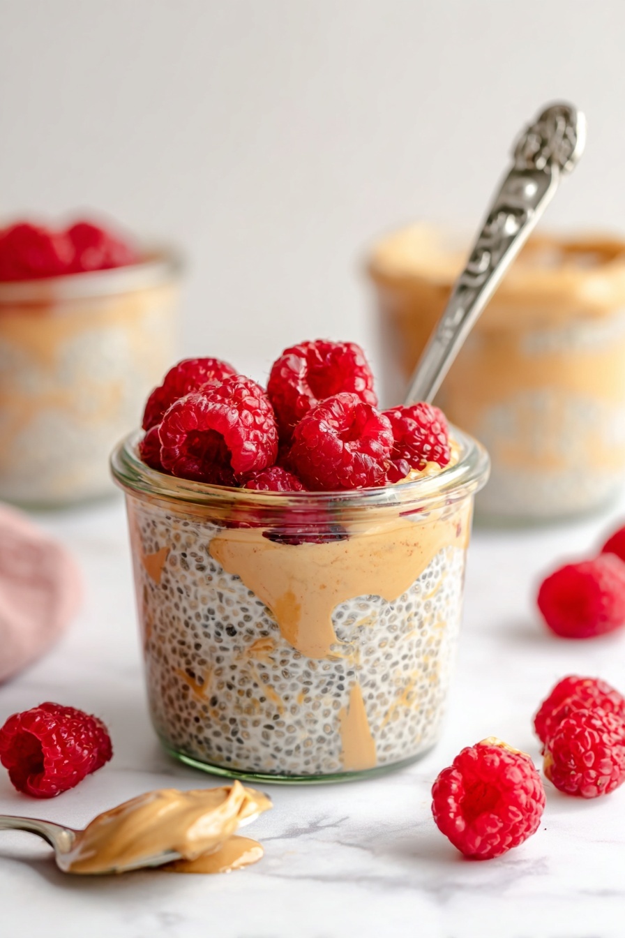 Peanut Butter Overnight Oats, healthy breakfast ideas, quick overnight oats, easy make-ahead breakfast, nutritious oatmeal recipes - A clear glass jar filled with three visible layers: the bottom layer is light beige chia seed pudding with black chia seeds mixed in, the middle layer shows swirls of creamy light brown peanut butter, and the top layer is a bright red cluster of fresh raspberries. A silver spoon with an ornate handle is placed inside the jar, leaning against the side with some pudding on it. Around the jar, scattered fresh raspberries add more red spots on a white marbled surface. In the background, more jars with similar contents and a silver spoon with a light brown spread are softly blurred. Photo taken with an iphone --ar 2:3 --v 7