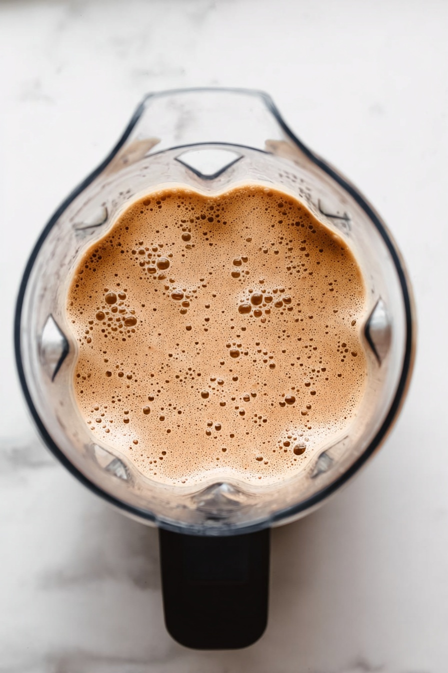 Vegan Chocolate Protein Shake, healthy vegan protein drinks, dairy-free chocolate shakes, plant-based protein smoothies, quick vegan breakfast recipes - The image shows a clear blender from above filled with a light brown, frothy mixture with many small bubbles on its surface. The blender has a black handle, and the background is a white marbled surface. photo taken with an iphone --ar 2:3 --v 7