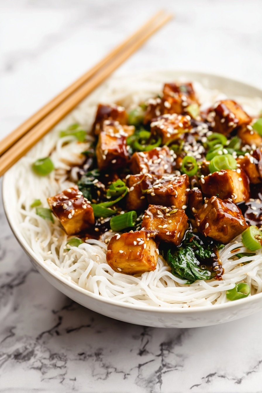 Quick One Pan Tofu Stir Fry, tofu stir fry recipe, easy tofu stir fry, healthy weeknight dinner, vegetarian stir fry - A white bowl is filled with thin white noodles forming the base layer. On top, there are medium-sized golden brown tofu cubes coated in a shiny dark brown sauce. Bright green chopped scallions and spinach leaves are mixed in with the tofu. White sesame seeds are sprinkled over the tofu pieces, adding texture and color contrast. Wooden chopsticks rest on the edge of the bowl over a white marbled surface in the background. Photo taken with an iphone --ar 2:3 --v 7