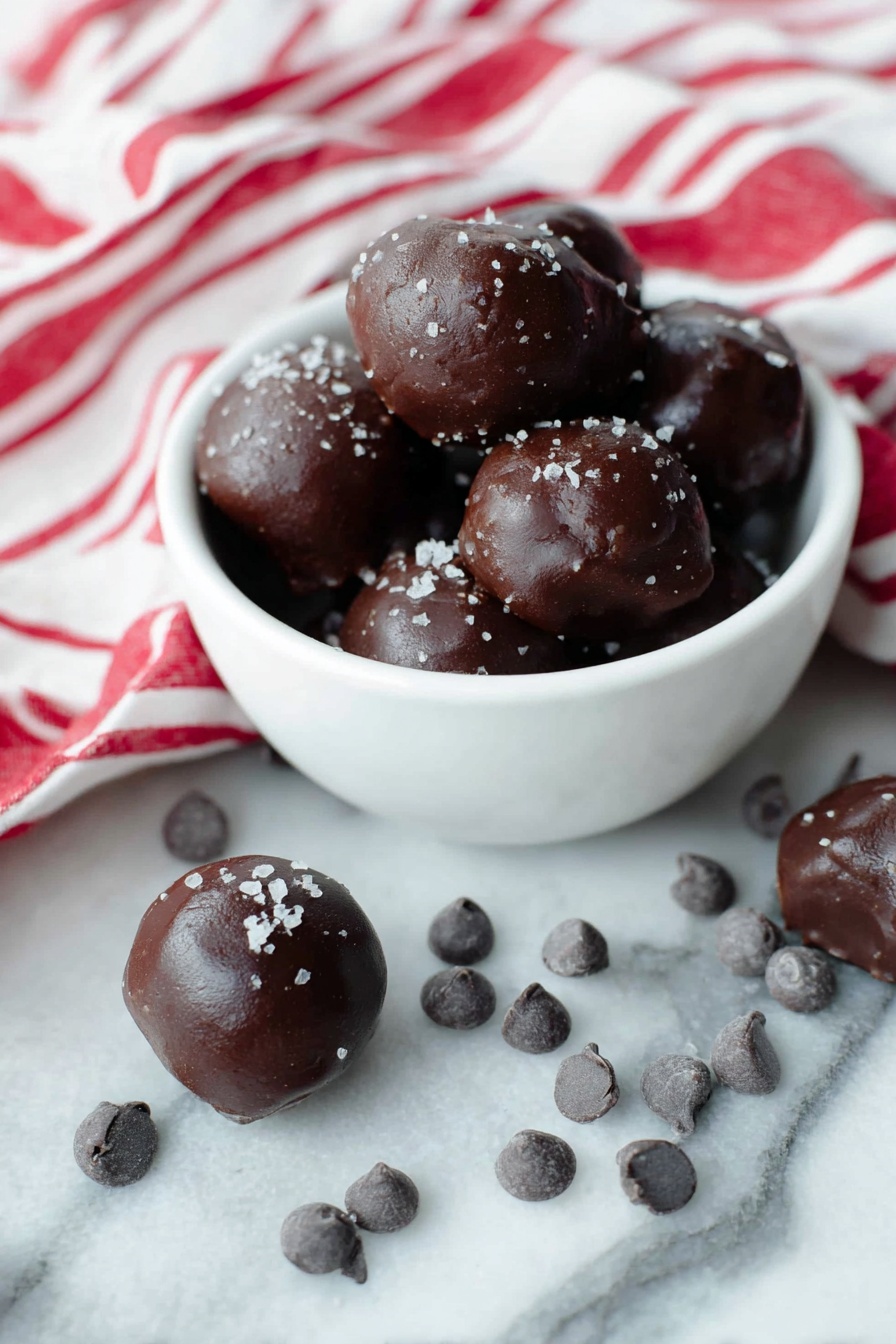 Cocoa Date Truffles, healthy chocolate truffles, vegan date bites, indulgent no-bake treats, natural sweet treats - A white bowl filled with several dark brown chocolate balls that have a smooth, shiny texture and are sprinkled lightly with coarse salt flakes on top. Around the bowl, more chocolate balls sit on a white marbled surface, some resting near small clusters of dark chocolate chips. A white cloth with bold red stripes is casually draped to the back and right side of the bowl, adding a bright contrast. The scene is bright and clean, showing the rich dark color and glossy texture of the chocolates clearly. Photo taken with an iphone --ar 2:3 --v 7