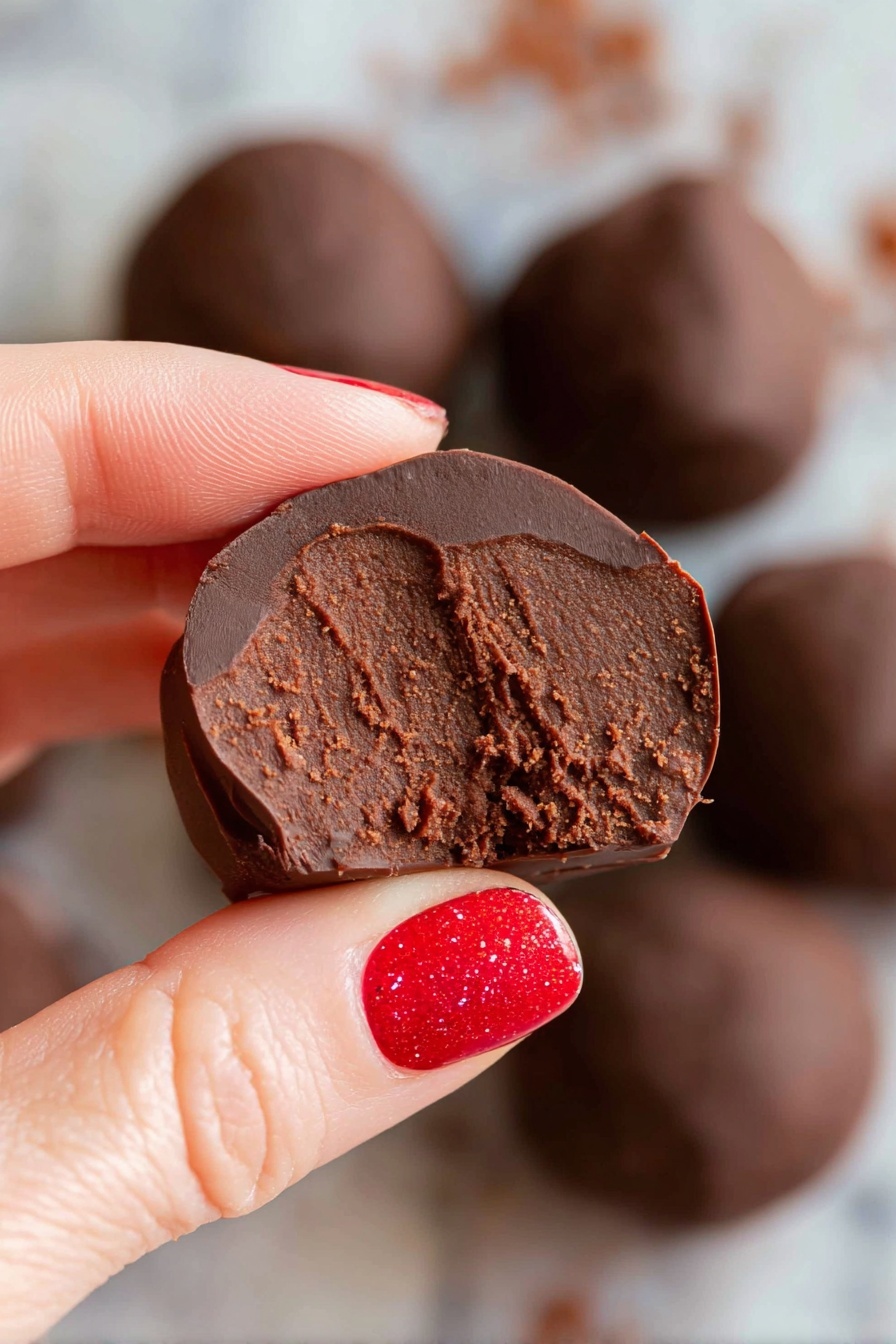 Healthy Chocolate Bean Snack Balls, healthy chocolate snacks, no-bake energy bites, nutritious chocolate treats, vegan chocolate snack balls - A round white bowl filled with several smooth, shiny dark chocolate balls stacked together. Two of the chocolate balls are cut in half and placed on top, revealing a dense, rich, and slightly grainy brown chocolate filling inside. The bowl sits on a white marbled surface, giving a clean and bright look. The chocolate balls have a glossy outer layer with some small wrinkles, showing a contrast between the shiny dark coating and textured inside. Photo taken with an iphone --ar 2:3 --v 7