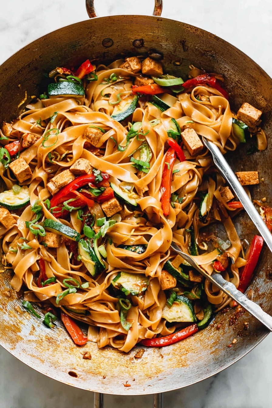 Drunken Noodles, Thai Drunken Noodles, Spicy Stir-Fry, Easy Noodles Recipe, Chicken and Vegetable Noodles - The image shows a metal wok filled with a colorful stir-fry dish. The base layer consists of wide, flat noodles that are light brown and glossy. Mixed through the noodles are pieces of lightly browned tofu cubes, thin strips of green bell peppers, slices of red bell peppers, and small chunks of zucchini with their green skin visible. Scattered over the top are thinly sliced green onions and bits of chopped herbs adding dots of bright green. The noodles and vegetables appear shiny from a light sauce coating them evenly, and the wok has a warm, well-used appearance with some browning on the sides. A pair of metal tongs rests inside the wok, ready to serve. The setting has a clean, white marbled texture surface. photo taken with an iphone --ar 2:3 --v 7