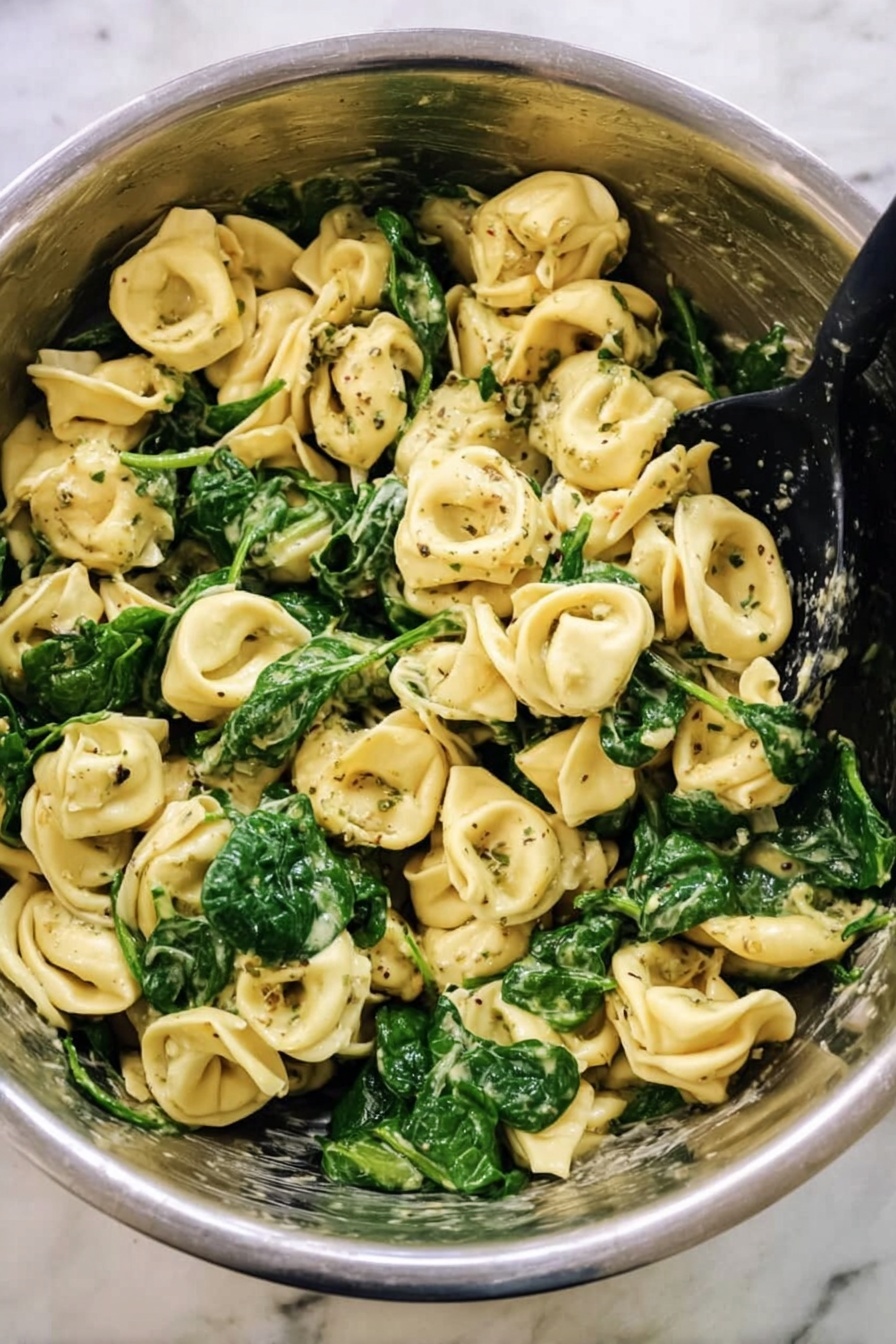 Creamy Pesto Tortellini, pesto tortellini recipe, quick pasta dinner, easy pesto pasta, creamy tortellini dinner - This image shows a big metal bowl filled with cooked tortellini pasta mixed with bright green spinach leaves. The pasta is light yellow with a soft texture, and some pieces have dark green herb specks on them. The spinach looks fresh and slightly wilted, spread evenly throughout the pasta. There is a black spoon resting inside the bowl, ready to serve. The bowl sits on a white marbled surface, giving a clean and simple background. Photo taken with an iphone --ar 2:3 --v 7
