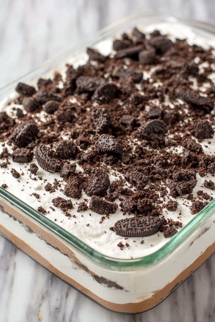 Red Velvet Poke Cake with Cheesecake Pudding, red velvet poke cake, cheesecake pudding dessert, easy red velvet cake, decadent poke cake - A clear glass rectangular dish holds a layered dessert with three visible layers. The bottom layer is a light brown creamy base, covered by a thick middle layer of smooth white cream. The top layer is sprinkled generously with roughly crushed dark chocolate sandwich cookies, scattered evenly across the white cream. The dish sits on a white marbled surface. photo taken with an iphone --ar 2:3 --v 7