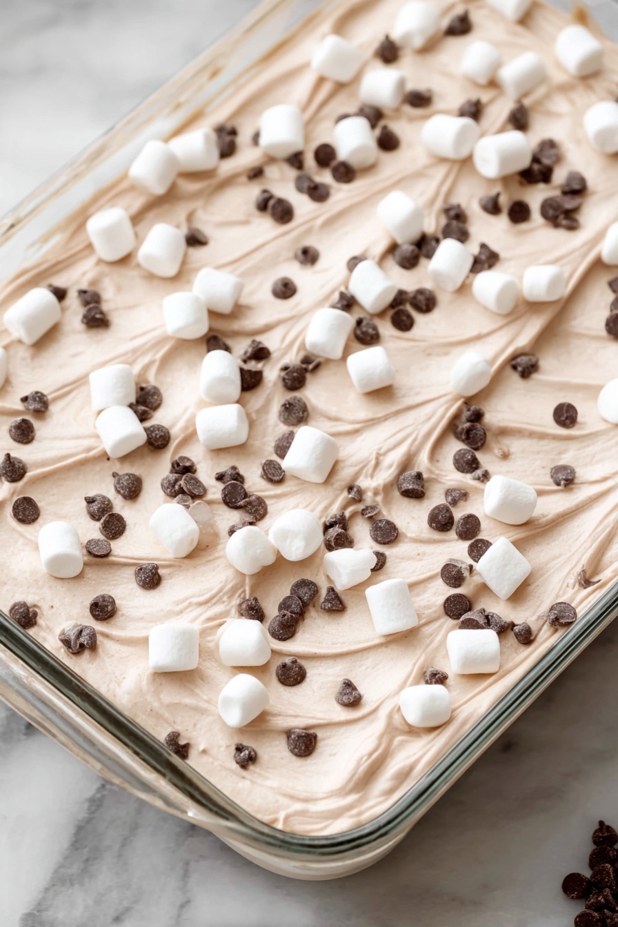 Hot Cocoa Poke Cake, chocolate poke cake with hot cocoa, marshmallow poke cake recipe, winter dessert poke cake, indulgent chocolate cake with marshmallow - A dessert in a clear rectangular glass dish with one thick layer of light brown creamy mousse or whipped topping, spread evenly with soft swirls on the surface. Scattered on top are many small white marshmallows along with a good amount of very tiny dark brown chocolate chips spread all over. The dish is placed on a white marbled surface. Photo taken with an iphone --ar 2:3 --v 7