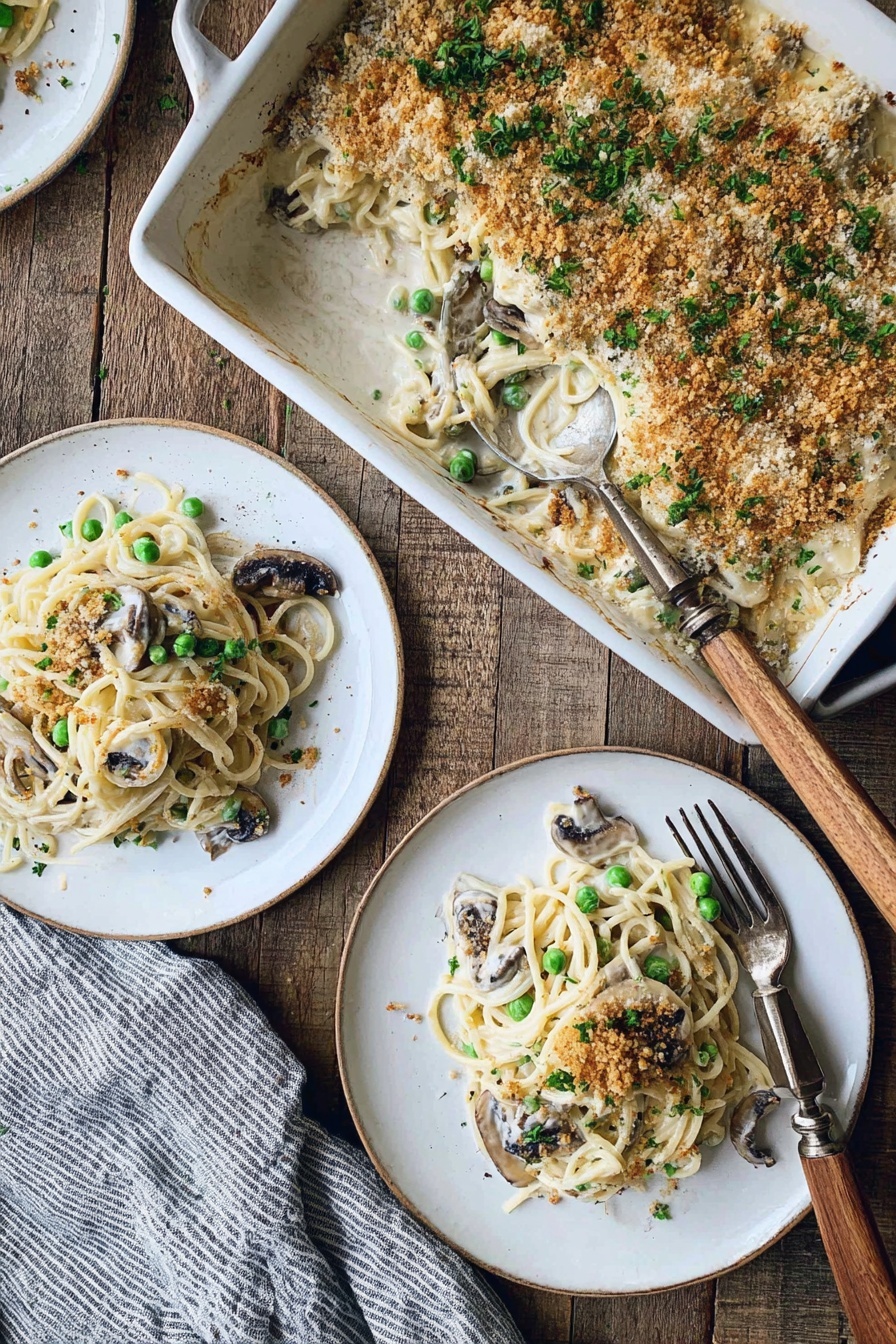 Creamy Turkey Tetrazzini, turkey pasta casserole, comforting turkey pasta dish, easy creamy turkey recipe, holiday leftover turkey dinner - The image shows a white rectangular baking dish filled with a creamy spaghetti casserole topped with a golden brown breadcrumb crust and sprinkled with chopped green herbs. Inside the dish, there are visible green peas and slices of dark mushrooms mixed with the creamy pasta. Two servings of the casserole have been plated separately on white round plates, each layered with creamy white sauce spaghetti, green peas, and mushrooms, and garnished with crumbs and herbs. A silver fork with a wooden handle rests on the main plate. The setting is on a rustic wooden table with a gray and white striped cloth nearby. Photo taken with an iphone --ar 2:3 --v 7