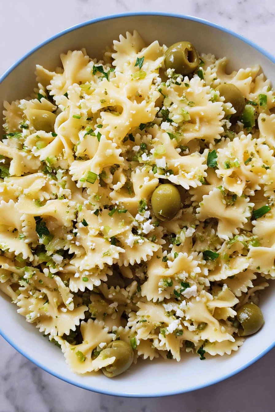 Dirty Martini Pasta Salad, olive pasta salad, blue cheese pasta salad, flavorful pasta salad, easy pasta salad recipes - A close-up view of a large white bowl filled with curly-edged pasta that is light yellow in color, mixed evenly with bright green sliced olives and sprinkled with small bits of light green herbs and crumbled white cheese. The pasta is separated nicely, showing the wavy edges and soft texture, while the green olives add contrast throughout. The bowl sits on a white marbled surface, creating a clean and fresh look. photo taken with an iphone --ar 2:3 --v 7