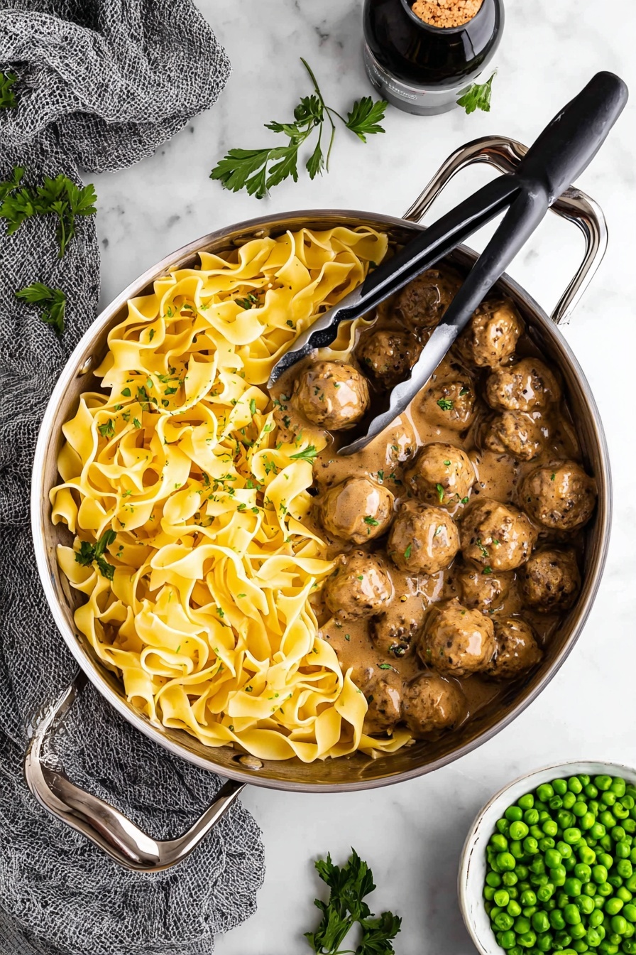 Swedish Meatballs with Creamy Gravy, Swedish meatballs, creamy gravy recipe, homemade Swedish meatballs, comfort food recipes - A large silver pan with tall handles holds two distinct layers inside: one side is filled with yellow wide pasta noodles twisted and folded over each other with a smooth, slightly shiny texture, while the other side has many round brown meatballs covered in a thick, creamy brown sauce with visible black pepper specks. Metal tongs with black grips rest on top of the pasta, slightly lifting some noodles. The pan is placed on a white marbled surface, surrounded by fresh green parsley leaves scattered around, a gray textured cloth to the left, a white bowl filled with bright green peas on the bottom right, and a dark bottle with cork inside at the top. Photo taken with an iphone --ar 2:3 --v 7