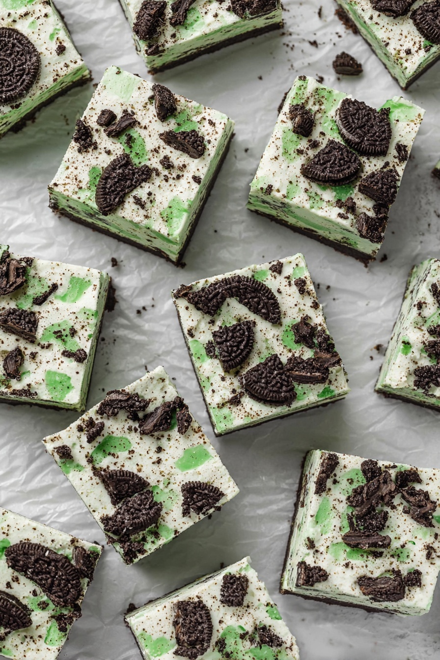 Mint Oreo Fudge, mint Oreo dessert, easy mint fudge, white chocolate fudge with Oreos, holiday mint fudge - The image shows many square pieces of dessert laid out on crumpled white paper over a white marbled surface. Each square has two visible layers: the bottom layer is a light creamy color with specks inside, and the top layer is a white cream with crushed dark chocolate cookies that have bright green filling scattered unevenly across the surface. The cookies add a rough texture on top, with pieces varying in size and shape. The squares are arranged closely but not touching each other, filling the image space evenly, and giving a look of a cool, creamy treat with a minty cookie crunch on top photo taken with an iphone --ar 2:3 --v 7
