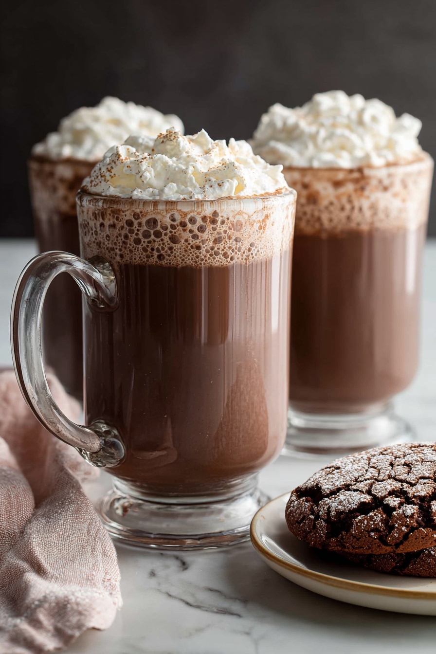 Homemade Hot Chocolate, cozy hot chocolate recipe, rich creamy hot chocolate, easy hot chocolate at home, best hot chocolate recipe - Two clear glass mugs filled with rich brown hot chocolate, topped with a thick layer of light white whipped cream that has a fluffy texture and soft peaks. The mugs have sturdy handles positioned on the left, and the hot chocolate shows slight bubbles near the edges under the whipped cream. In the foreground, on the right side, there is a white plate with a beige rim holding two cracked chocolate cookies with a powdery surface. A soft pink and white cloth napkin is casually placed on the left side near the front mug. All items sit on a smooth white marbled surface with a dark, blurred background behind them. Photo taken with an iphone --ar 2:3 --v 7