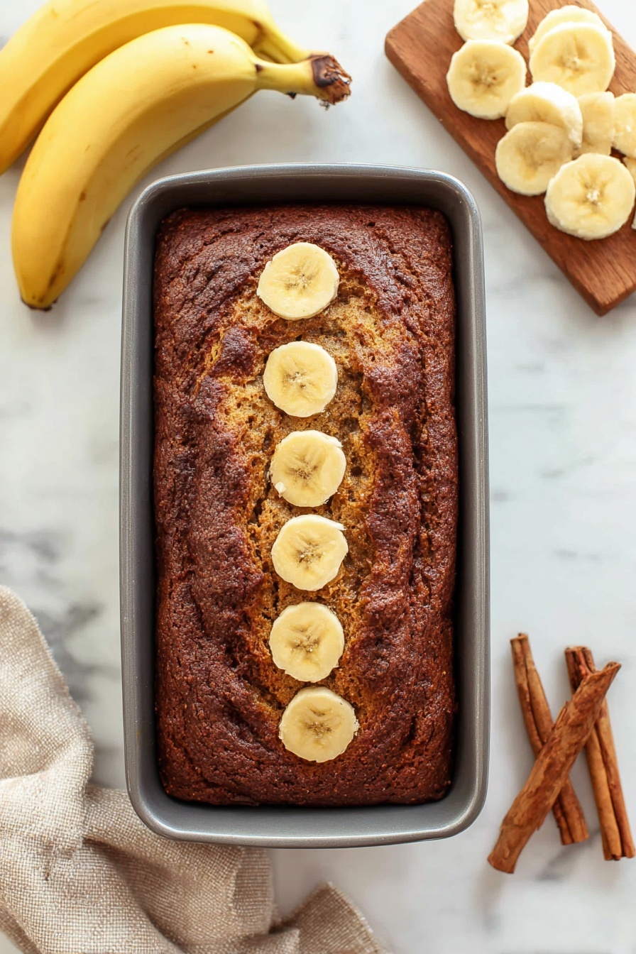 Cinnamon Banana Bread with Swirl, banana bread recipe with cinnamon swirl, moist banana bread with cinnamon, easy cinnamon banana bread, homemade cinnamon banana loaf - A thick rectangular loaf of dark brown banana bread rests in a grey baking pan, with five round banana slices lined up in the center top of the bread. To the left, two whole yellow bananas lay curved on a white marbled surface. To the right, a wooden board holds several light yellow banana slices arranged in overlapping circles. At the bottom right corner, three brown cinnamon sticks rest on the white marbled surface, and a beige cloth is partially visible at the bottom left corner. photo taken with an iphone --ar 2:3 --v 7