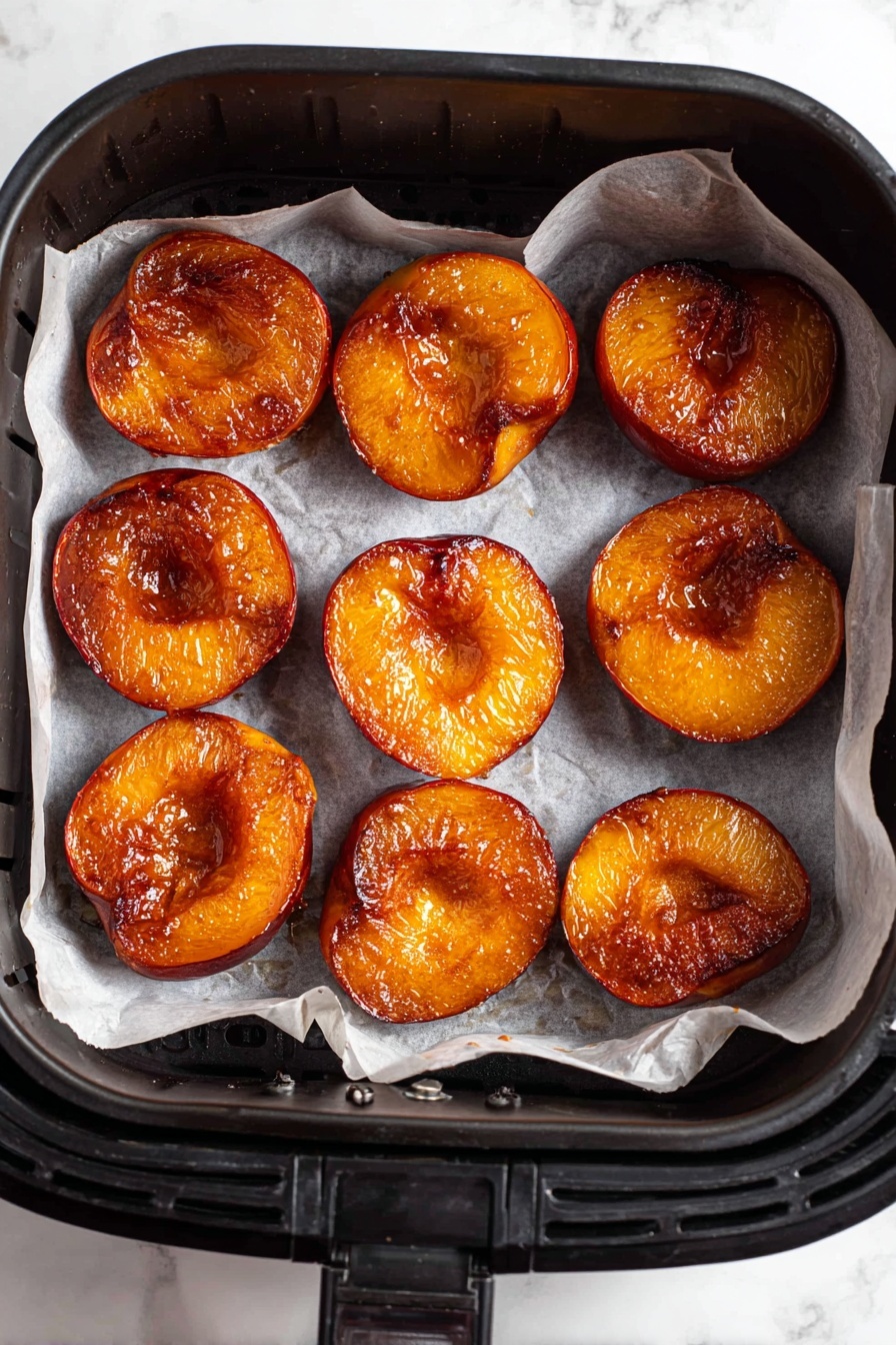 Air Fryer Peach Halves with Cinnamon, healthy peach dessert, quick fruit dessert recipe, caramelized peaches in air fryer, cinnamon peach halves - The image shows eight halved peaches placed inside a black air fryer basket lined with white parchment paper. Each peach half has a rich reddish-orange color with a shiny, caramelized texture, showing a light glaze over the surface. The peaches are arranged in two uneven rows, filling most of the basket's flat base. The background visible outside the air fryer is a white marbled texture. photo taken with an iphone --ar 2:3 --v 7